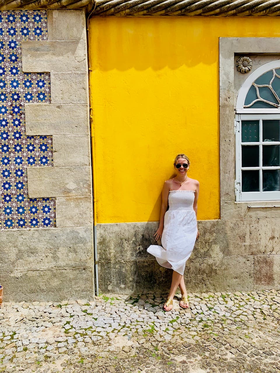 Allison wearing a white dress and posing in front of a vibrant yellow wall near a white-framed window and stone wall.