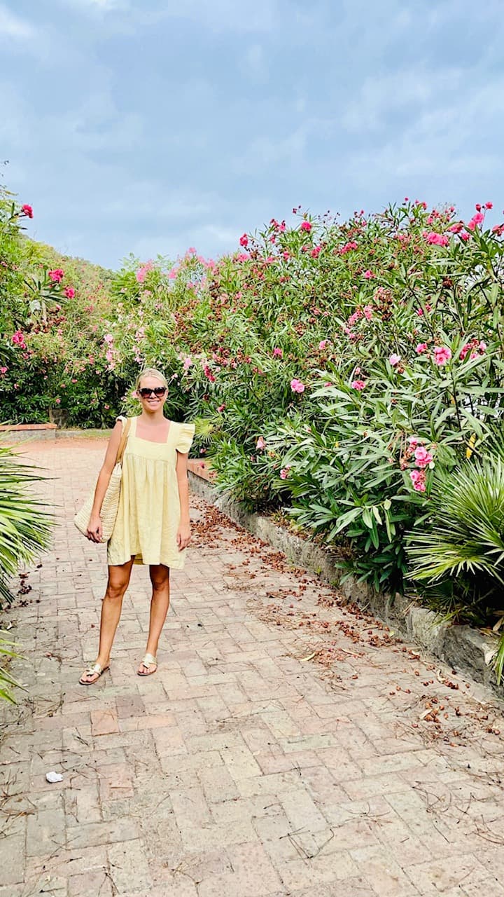 Allison walking down a stone path with a yellow dress on and pink floral filled bushes in the surrounding areas.