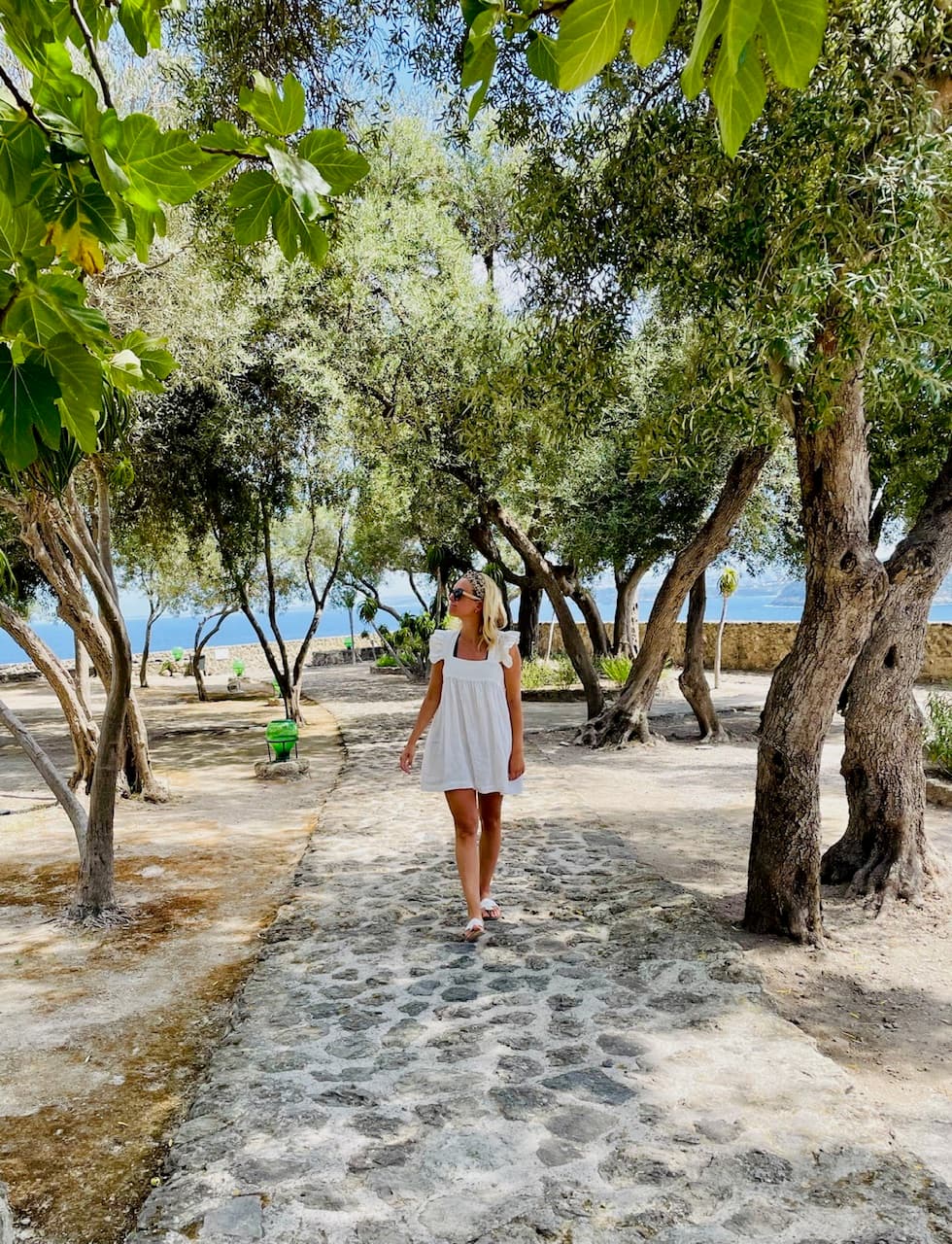 Allison walking down a stone path while wearing a white dress outside. There are trees and a body of water in the background and surrounding areas.