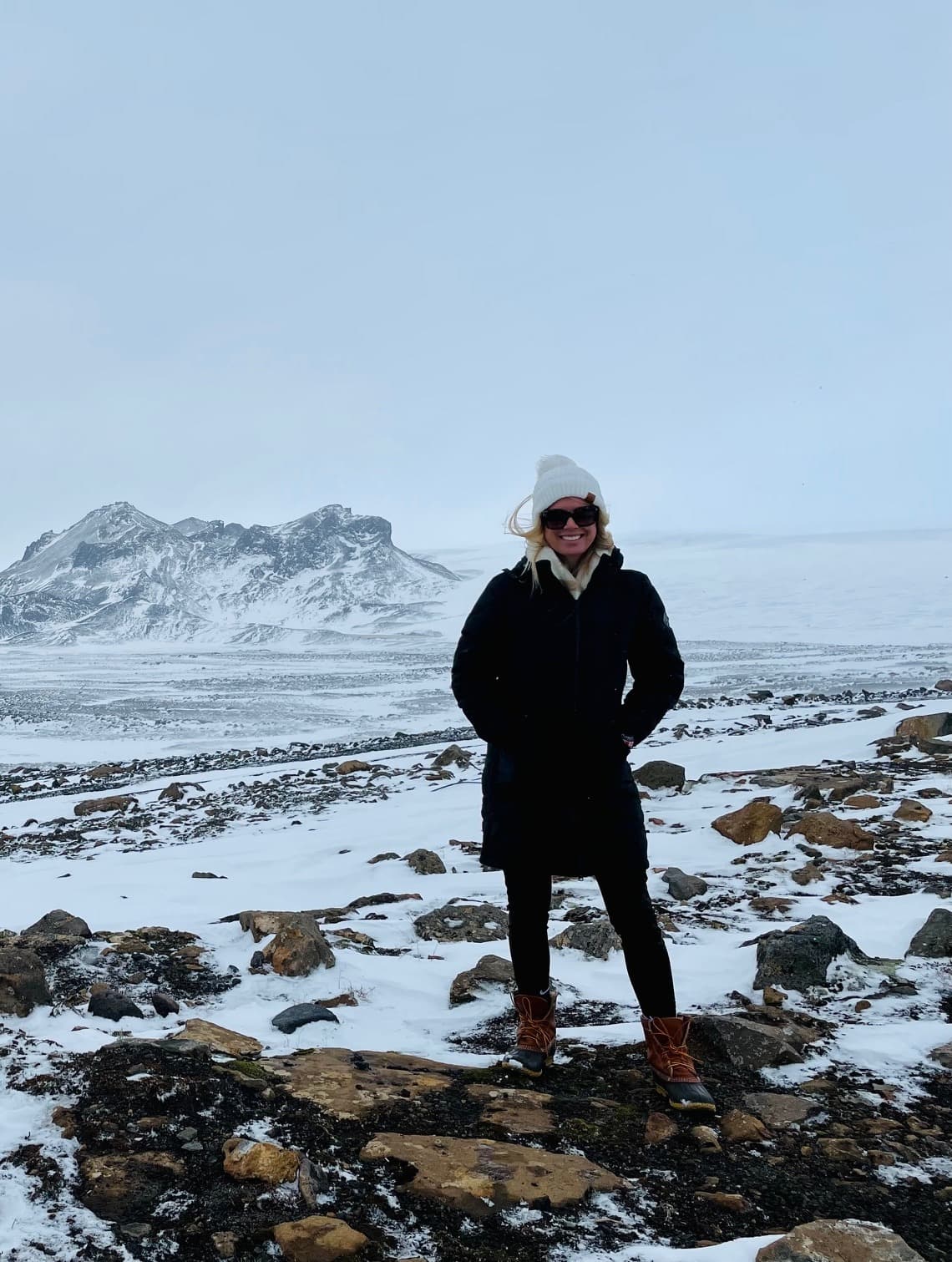 A woman posing outside in front of snowy terrain and a rocky, rugged mountain.