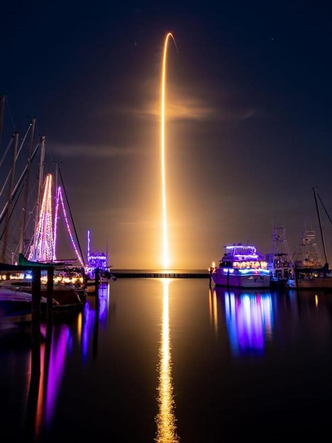 A rocket launch in the dark over a harbor with colorfully lit boats in the foreground.
