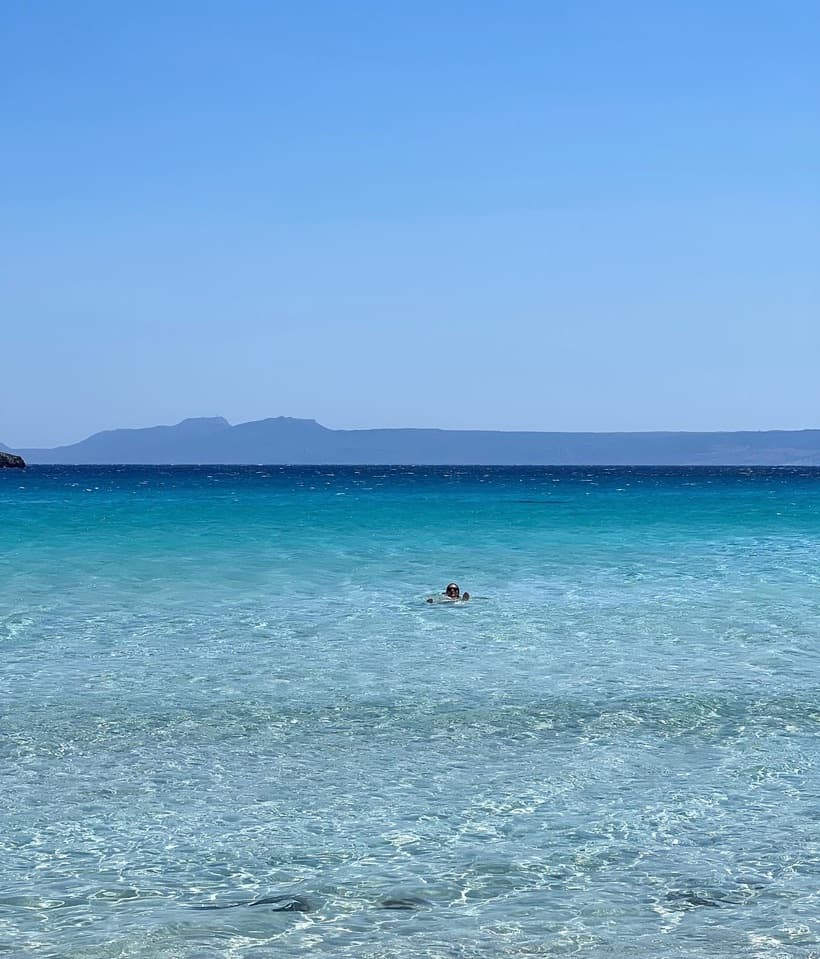 A beautiful view of crystal-clear sea water. There is a person swimming and a strip of land in the distance.