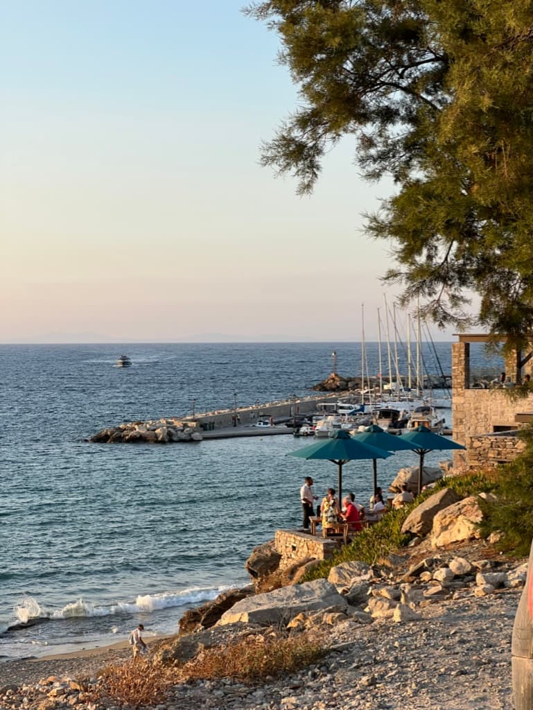 A beautiful view of a beach with people standing near blue umbrellas, boats docked and trees.