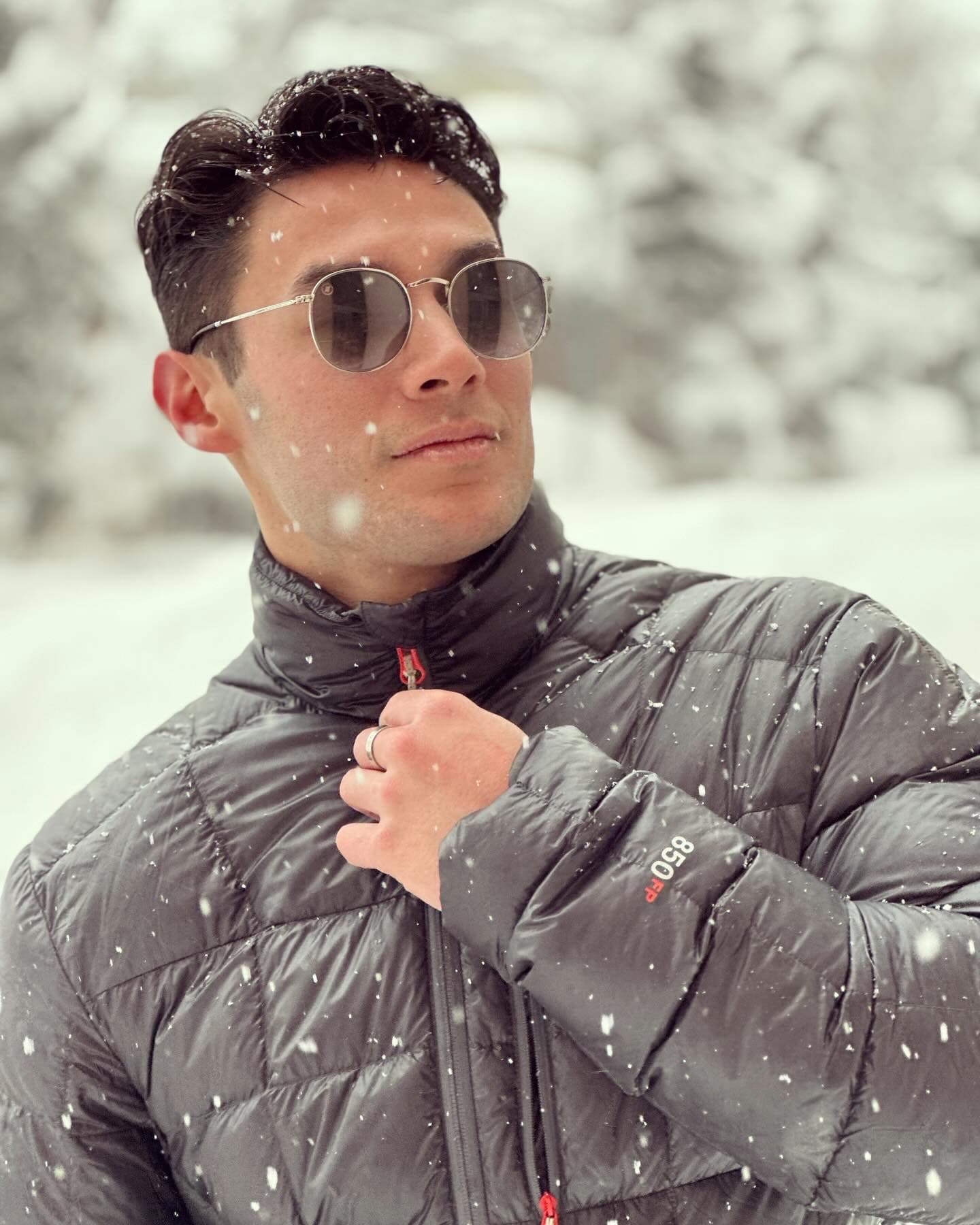 A man tugging on his coat zipper and wearing sunglasses while posing for a photo outside as it snows.