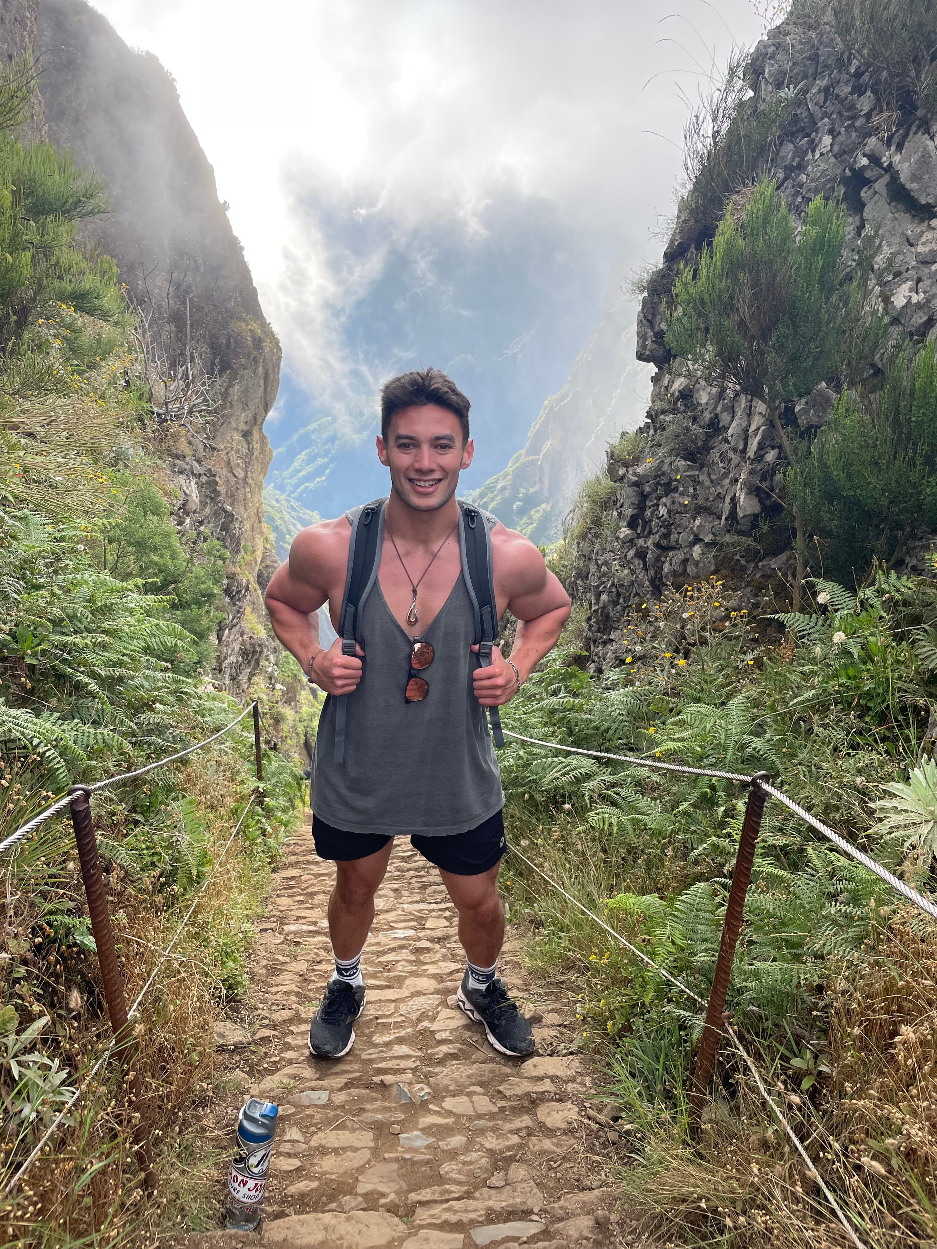 A man posing for a photo in the middle of a hiking trail. There is brush, large rocky cliffs and fog in the surrounding areas.