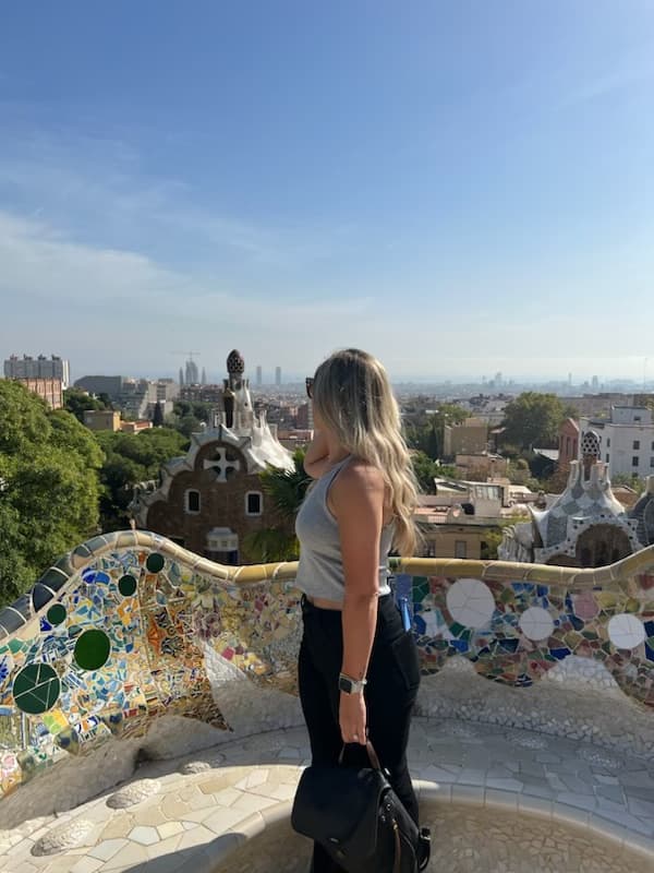 A woman wearing a grey top and black pants standing next to a stone bench with mosaic tiling. There is a view of tree tops and a city scape.