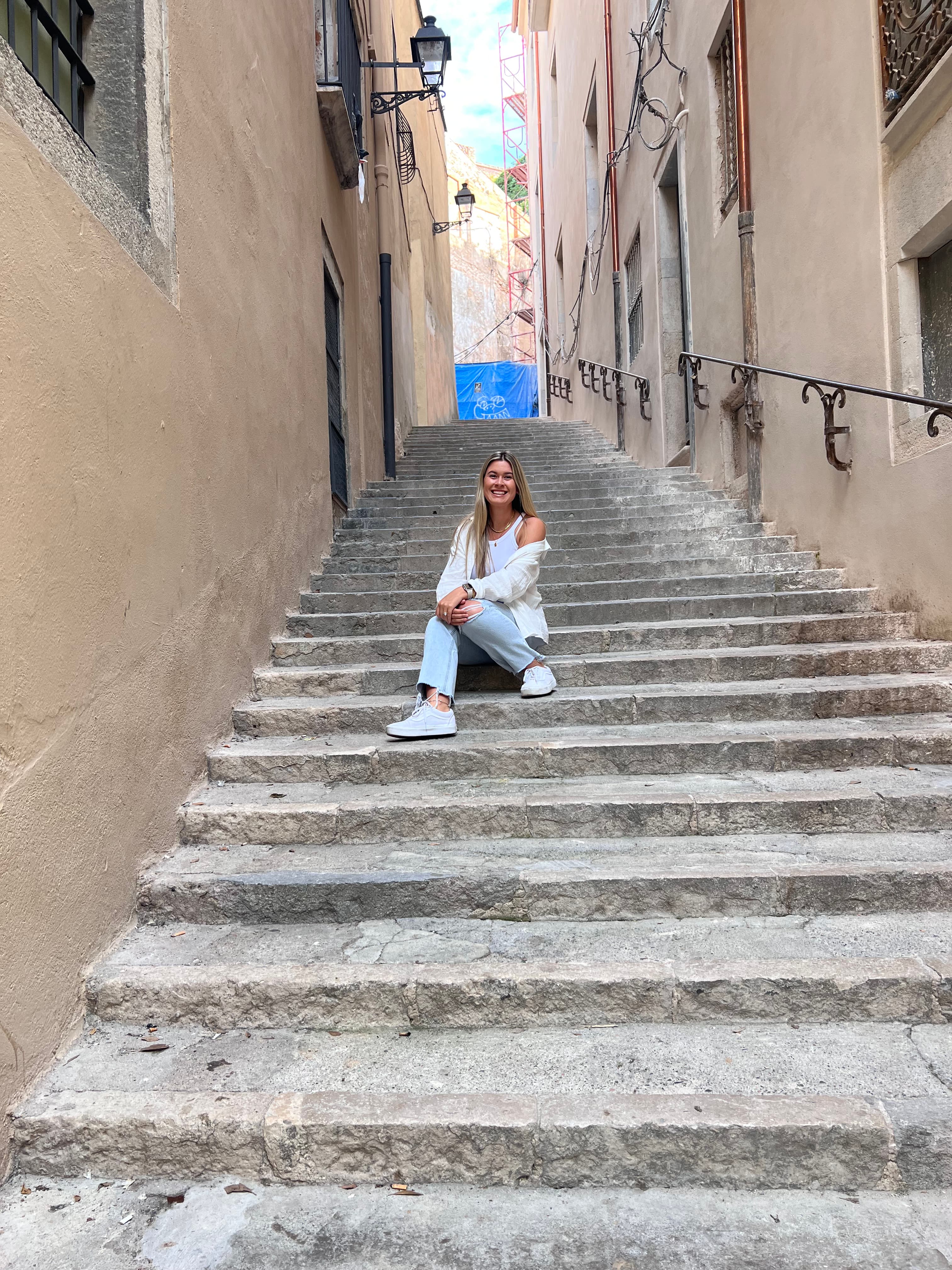 A woman wearing a white top and blue jeans on stone steps. There are stone walls and a metal railing to her right.