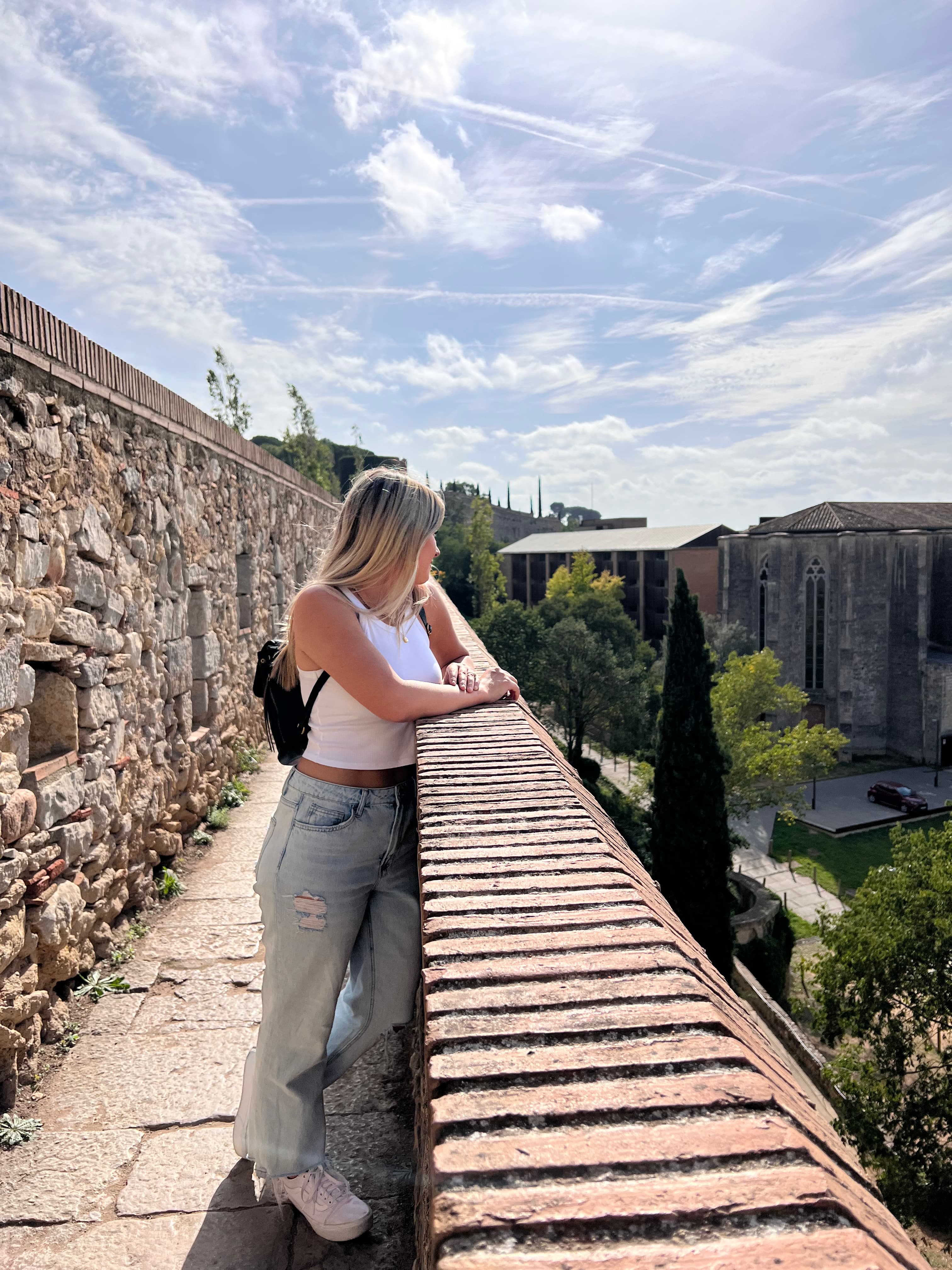 A woman wearing a white top and grey pants while leaning against a stone ledge. She is looking out towards a view of trees and a city scape. There is also a stone wall behind her.