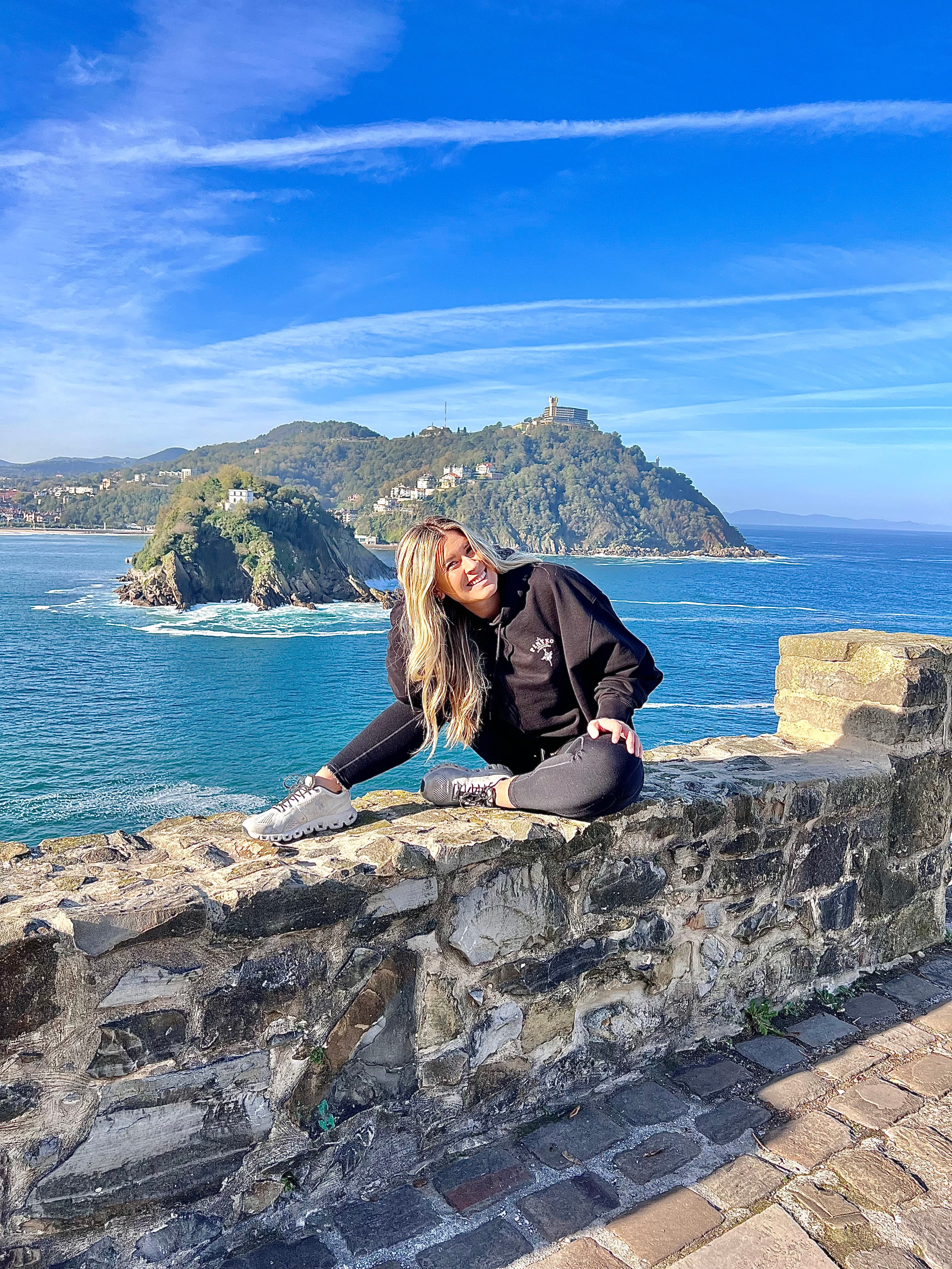 A woman wearing a black top and pants posing on top of a brick barricade with a view of vibrant blue water and large rock formations in the background.