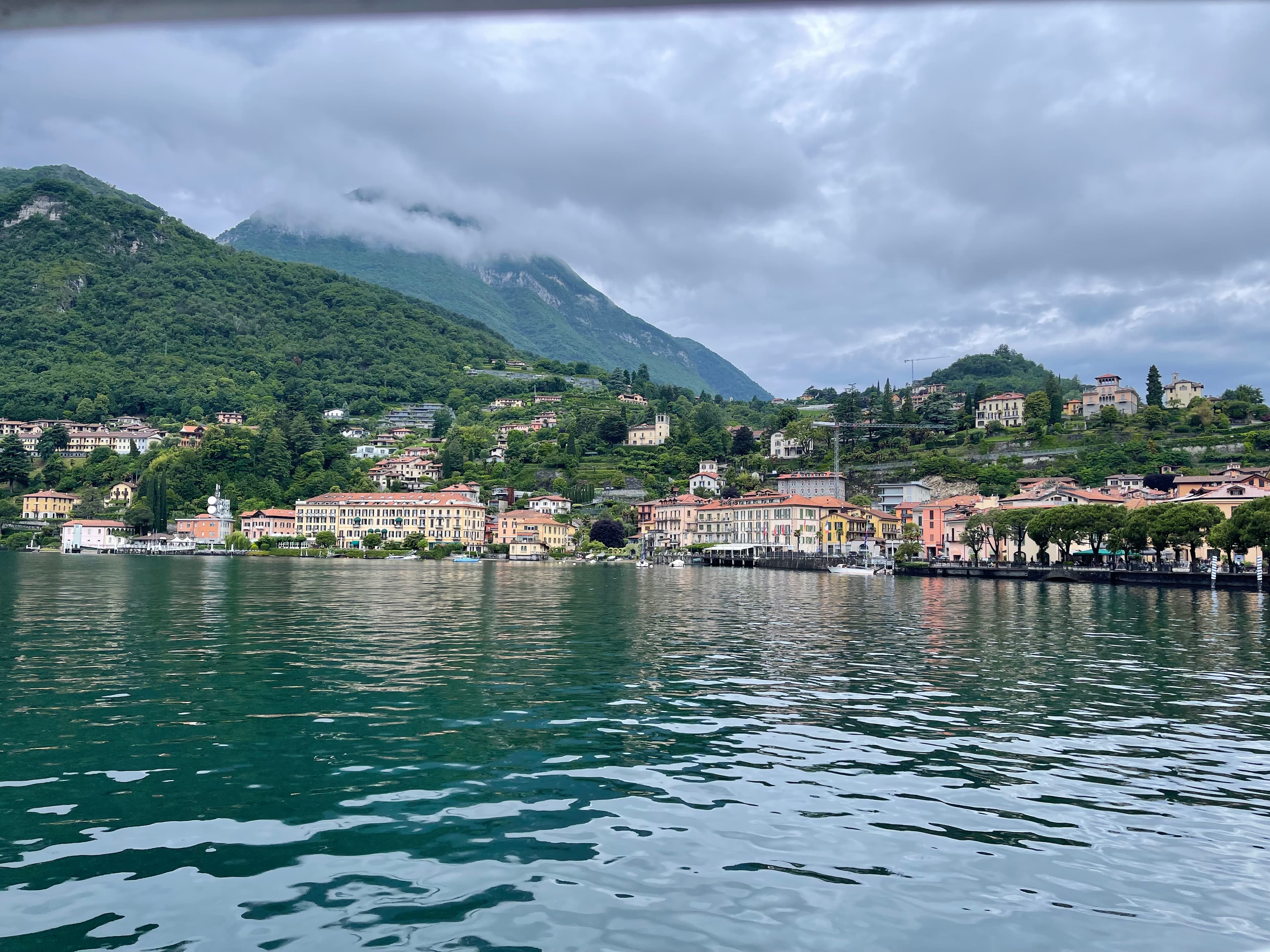 Red-roofed houses along a lake with green hills in the background on a cloudy day.