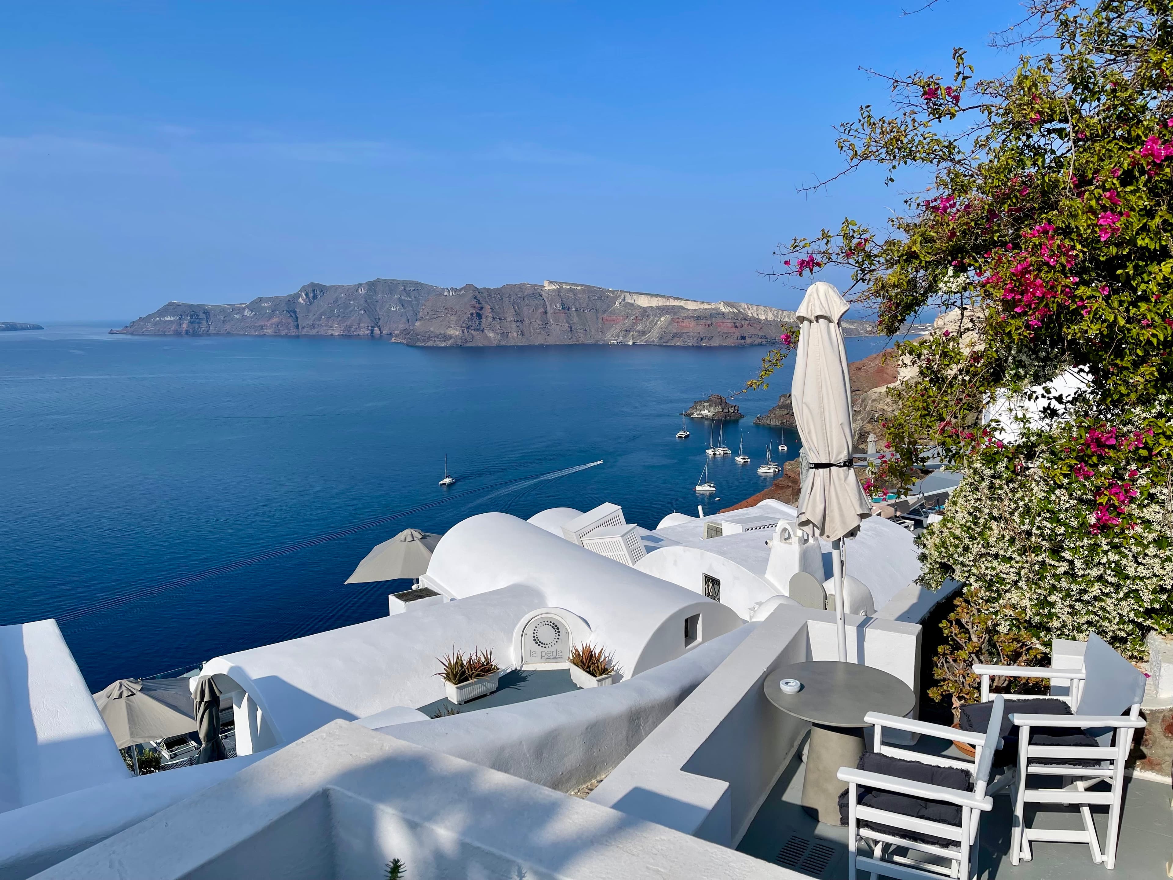 Santorini, with a small patio and chairs overlooking white cliffside houses and the ocean below, with an island in the distance.