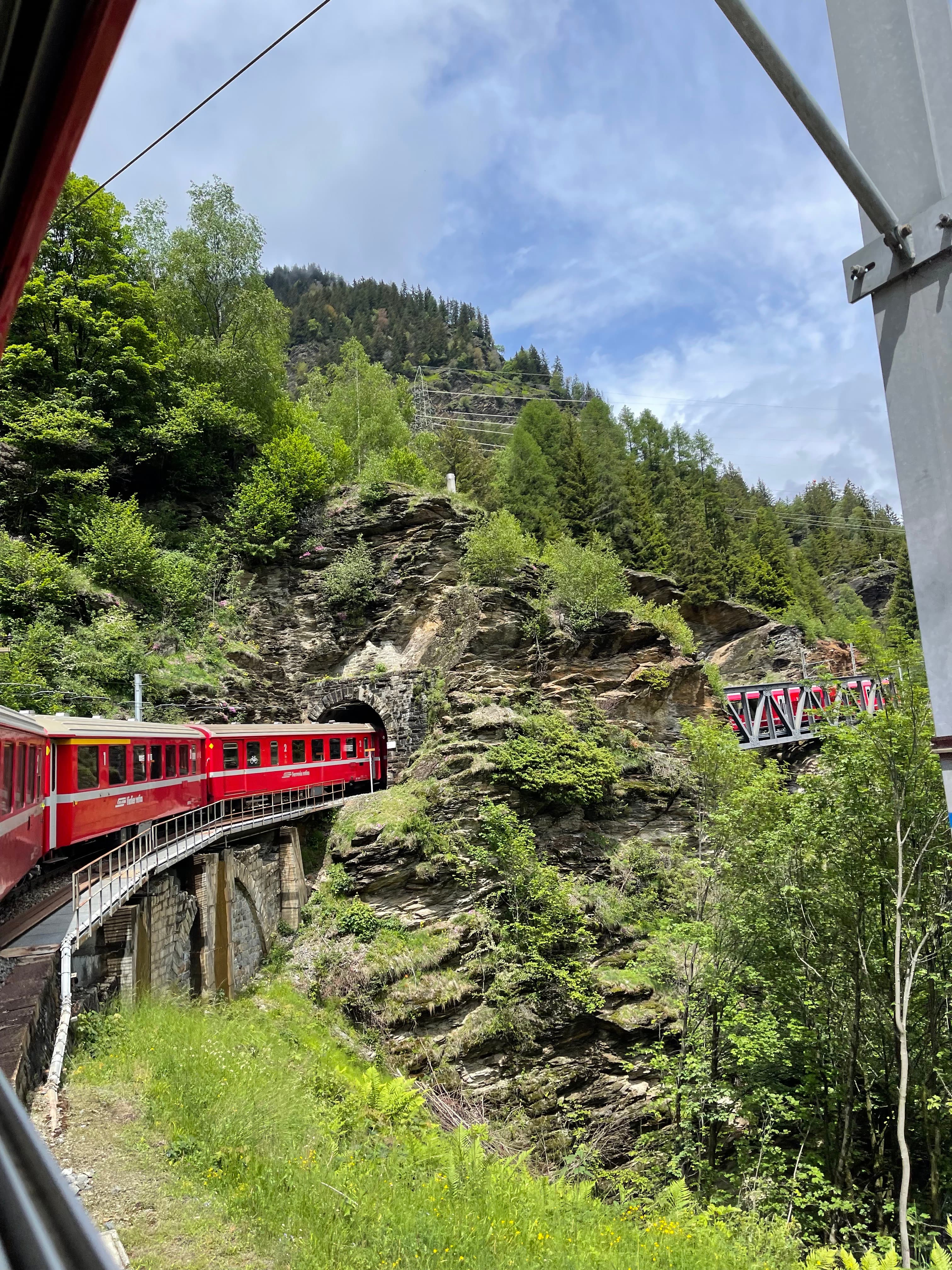 A red train on an elevated mountainside bridge traveling through a tunnel.