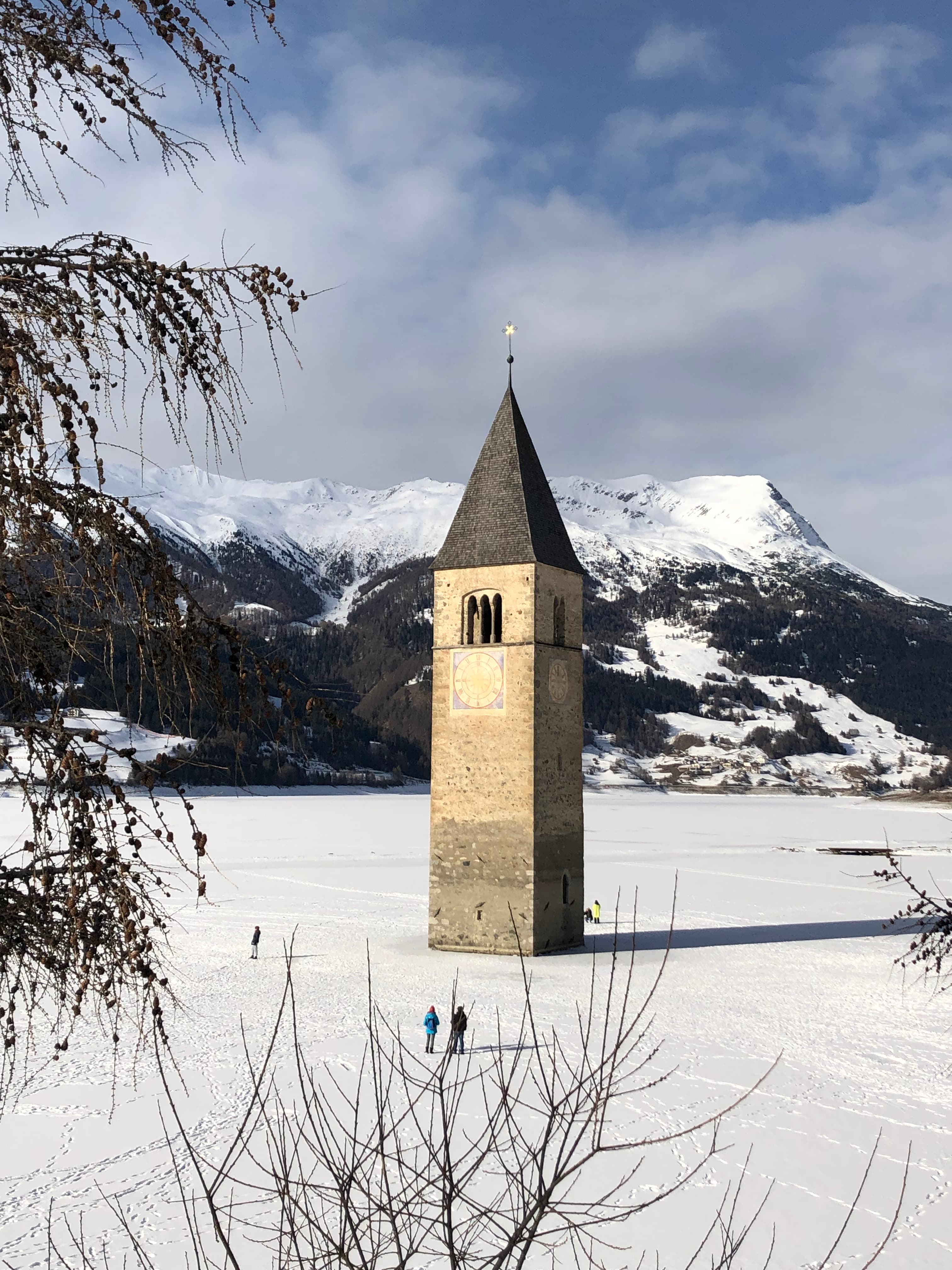 A stone tower in the middle of a snowy field with people walking, dried up trees and snowy mountains in the background.