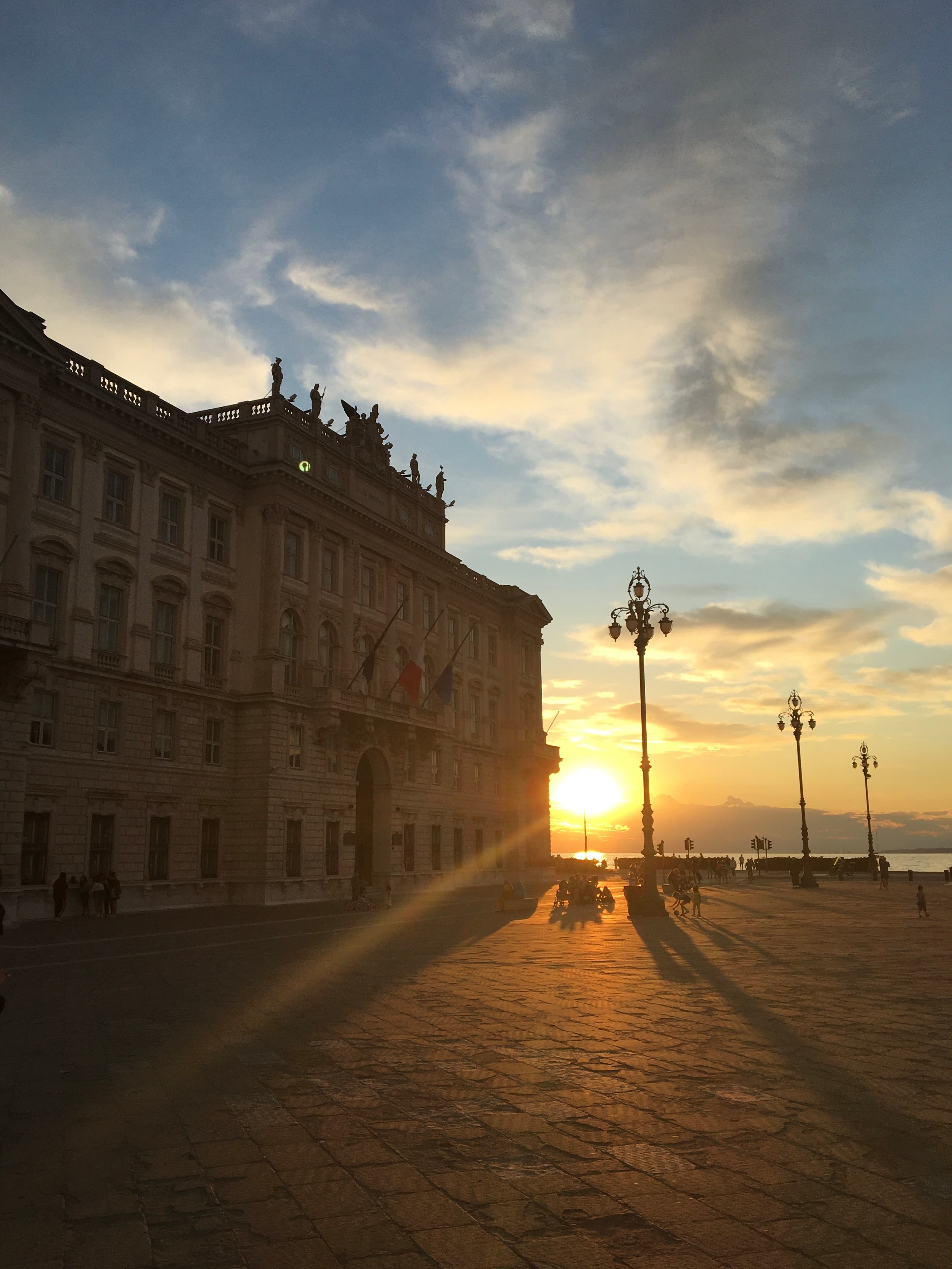 A European city street with stone buildings on the left, light posts on the right, a brick road and a beaming gold sunset shining through in the middle.
