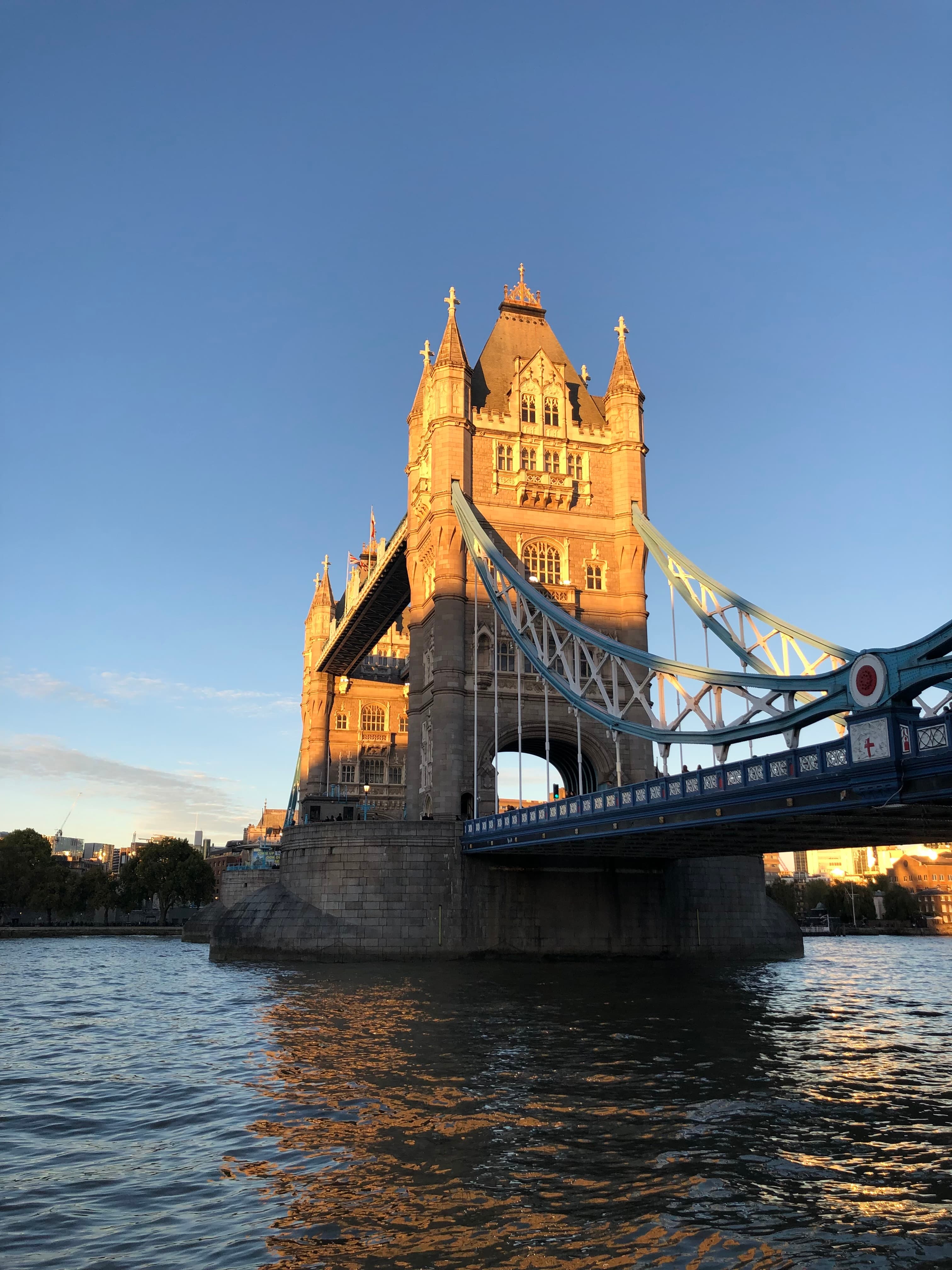 A view of a bridge in London with a large stone tower hanging over a reflective river. The sun is shining onto the tower.