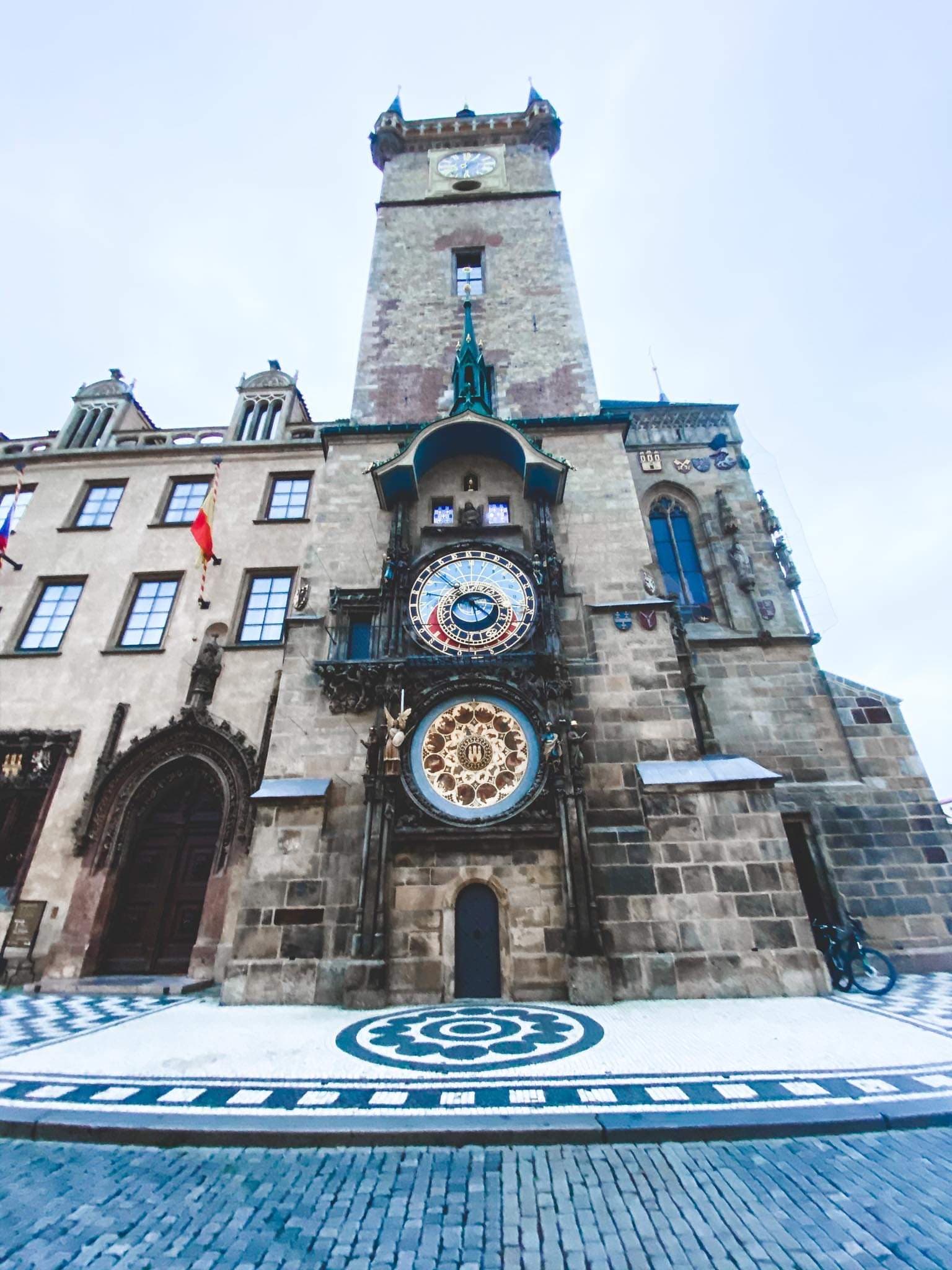 An old clock tower with stonework, windows, flags and a brick road.