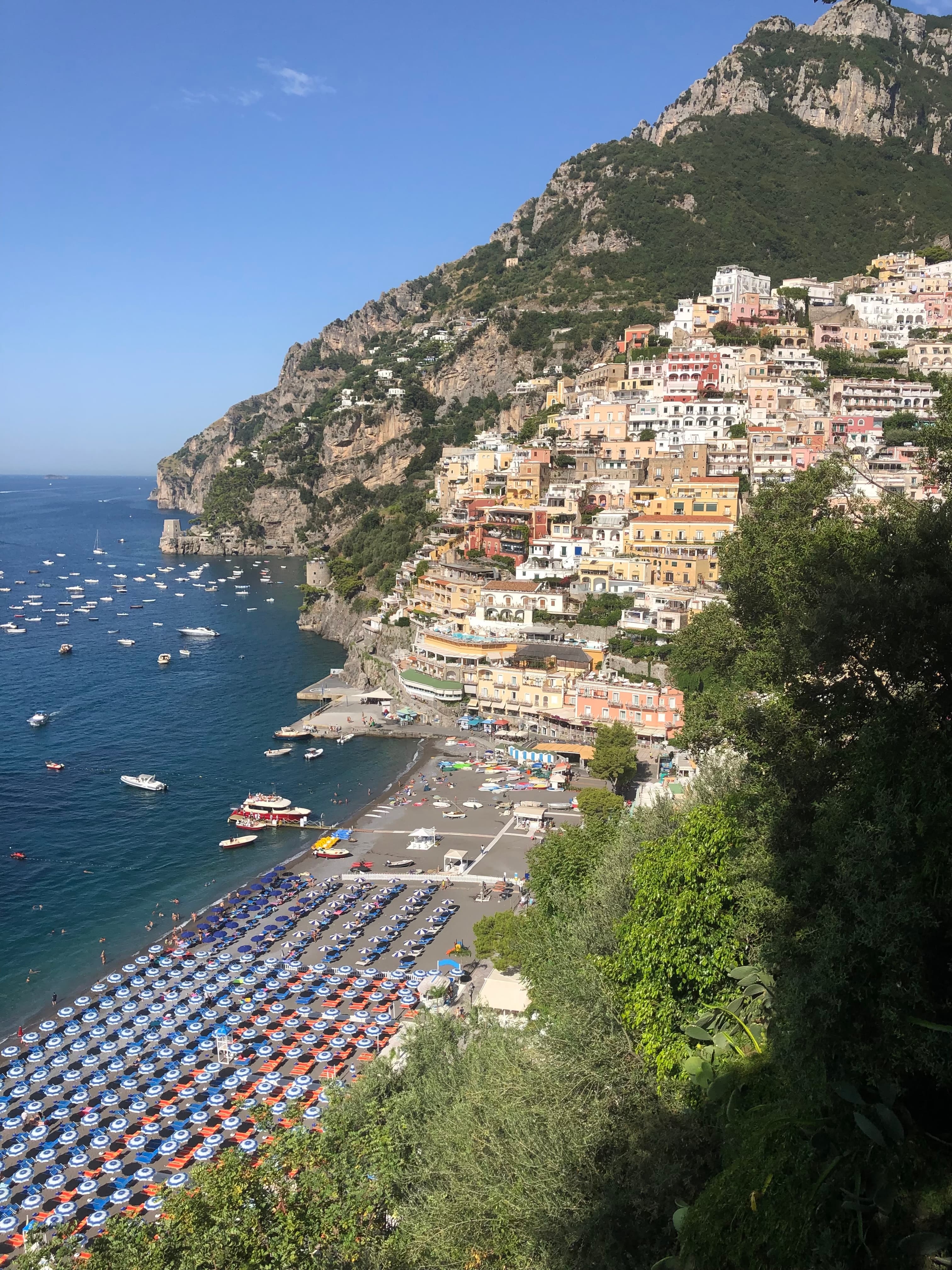 A beautiful view of an Italian beach with a uniform array of beach umbrellas, boats in the water and a colorful coastal town nestled into the green and rocky mountain.