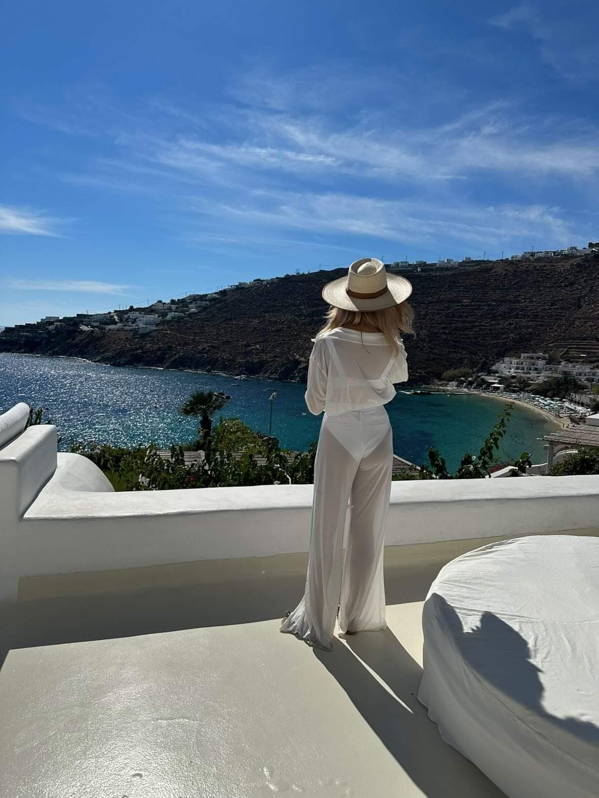 Beatrice in a linen coverup and straw sun hat standing on a patio facing the water.