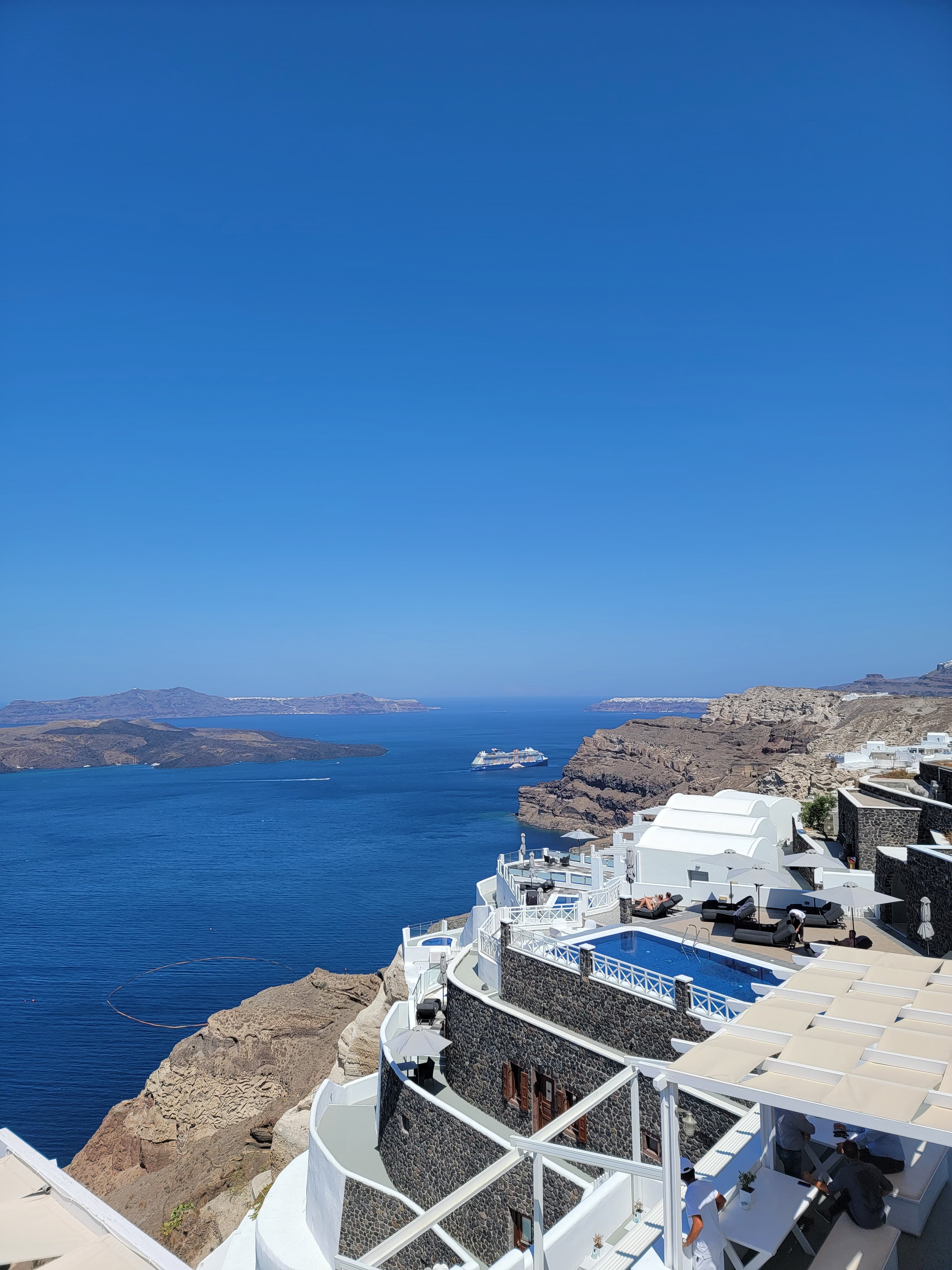 A beautiful view of stone, white terraces with swimming pools and people overlooking the rocky cliffs and vast blue sea with mountains in the distance.