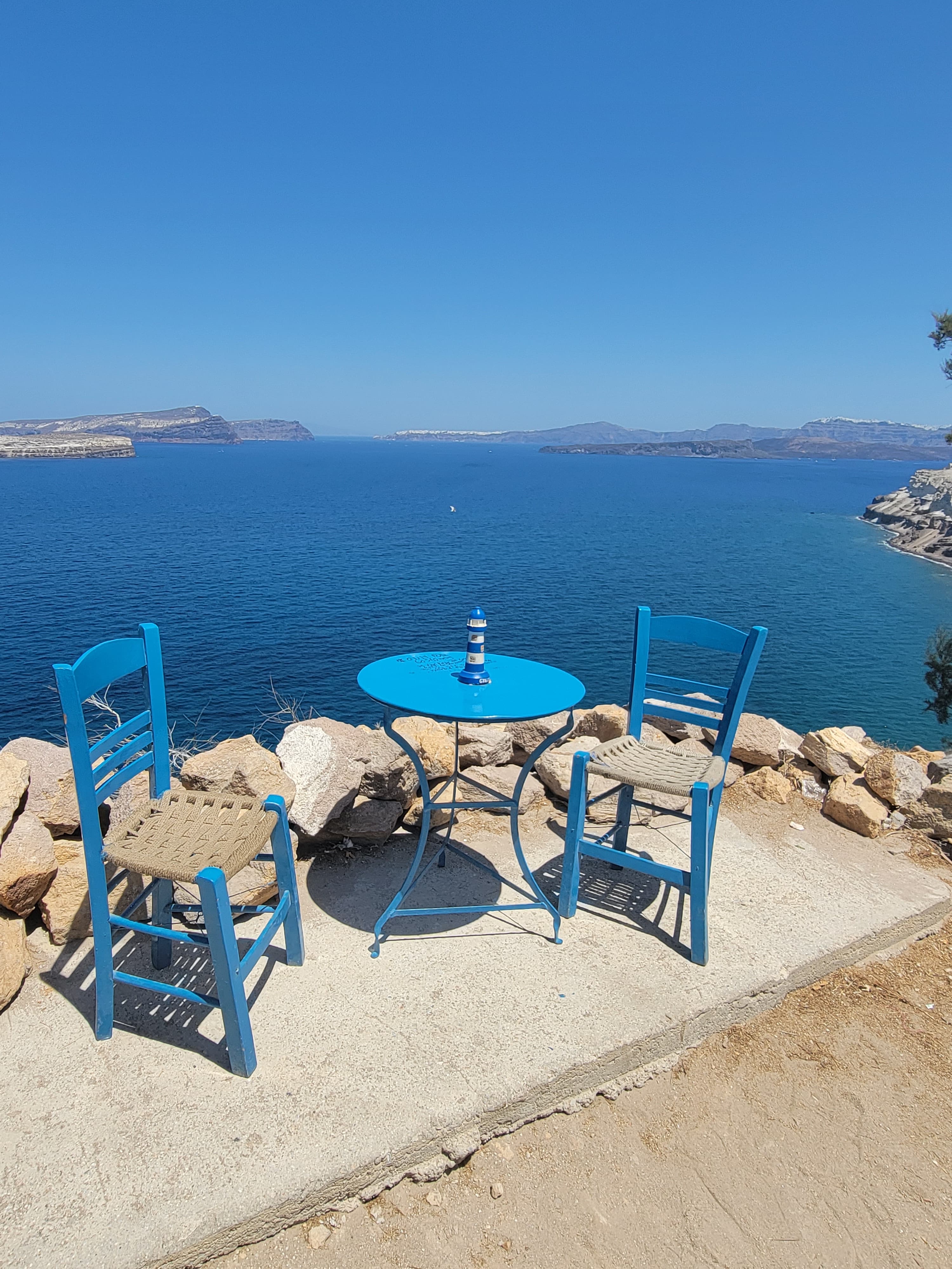 A blue table and two matching blue chairs positioned in an alcove that looks out to a view of the vast, blue sea with mountains in the far distance.
