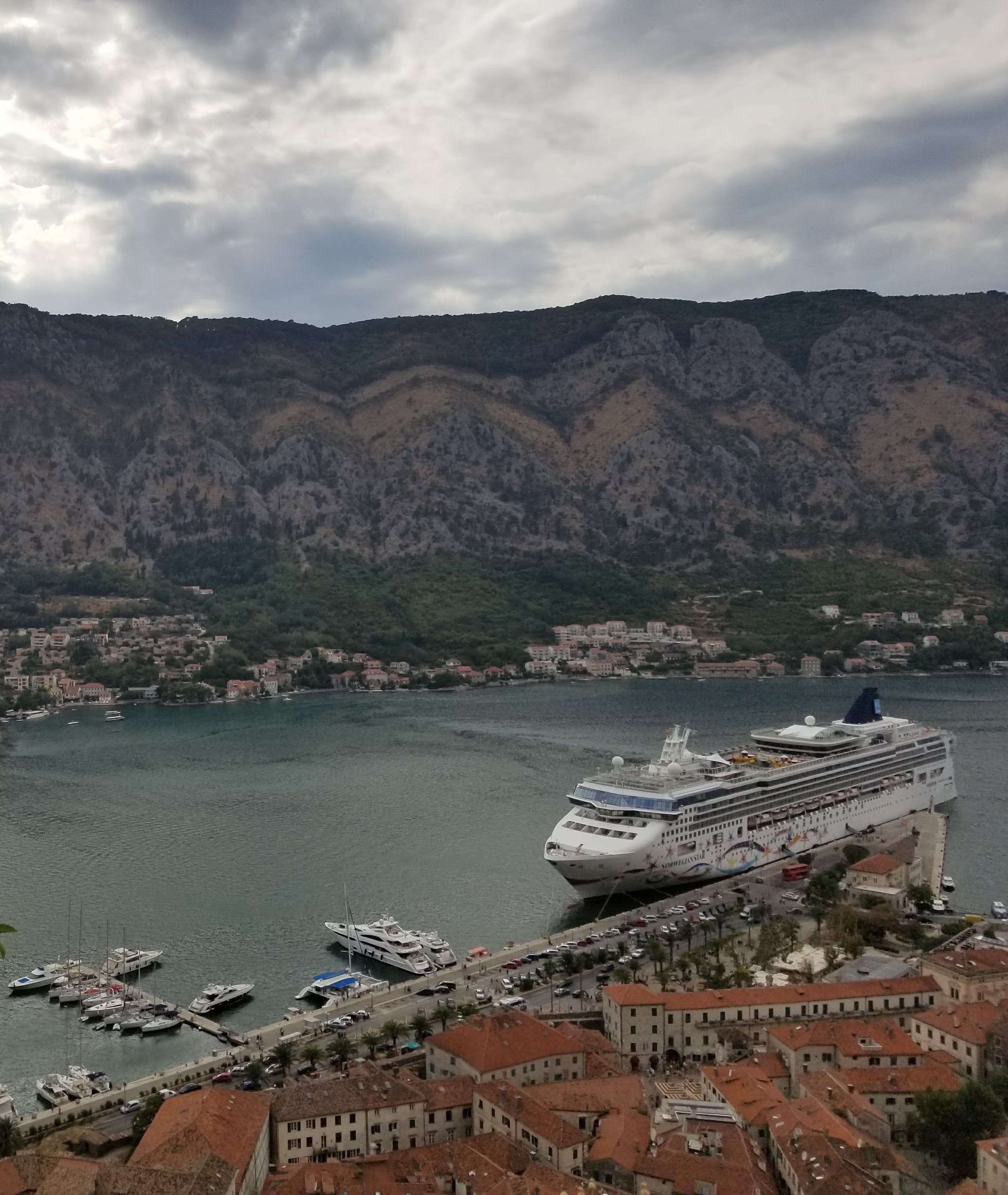 An aerial view of a cruise ship on top of a body of water surrounded by rocky mountains, coastal towns and red roofed buildings.