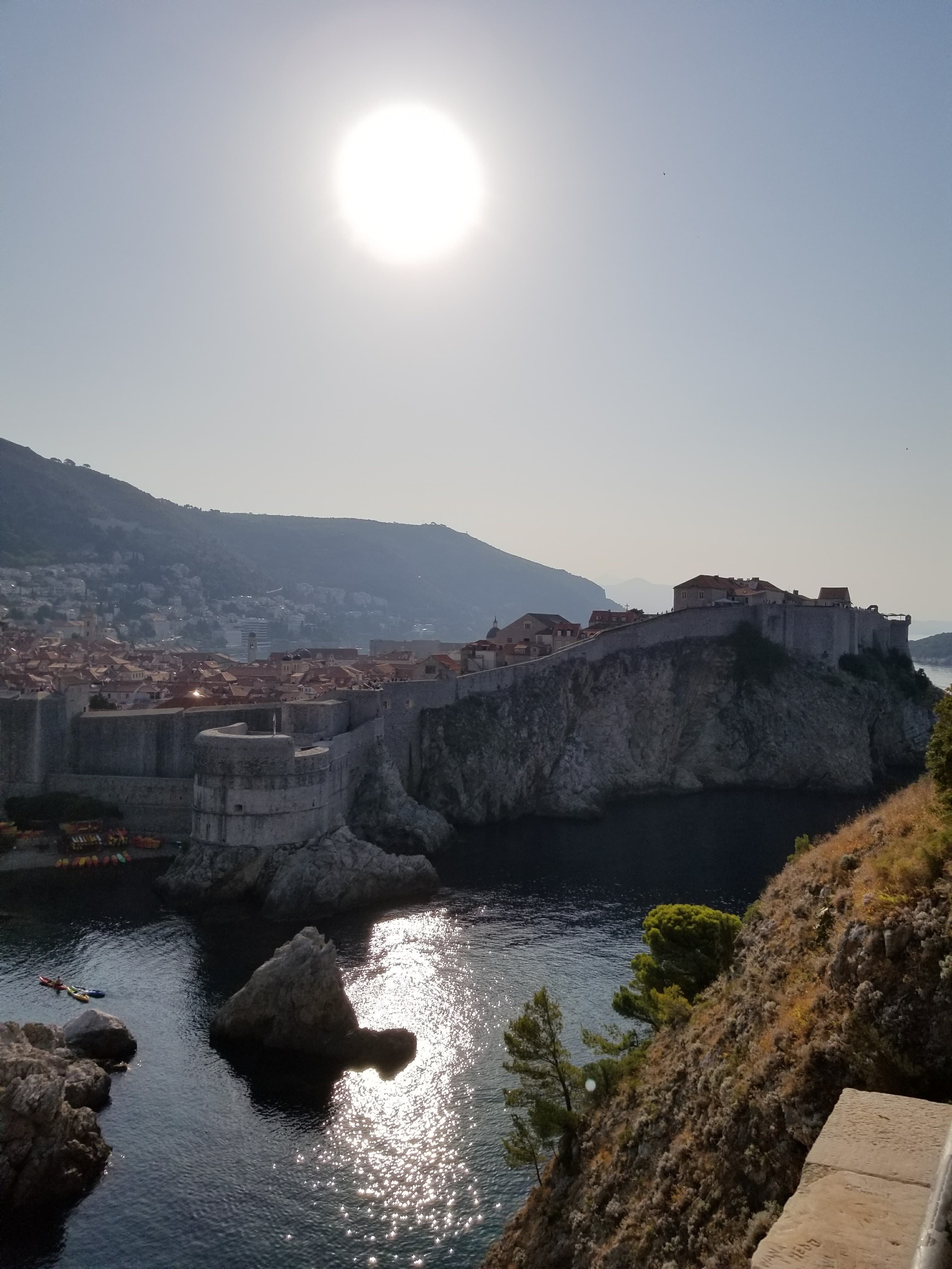 A beautiful view of Fort Lovrijena, surrounded by rocky cliffs, glistening water, shrubs and the bright sun.
