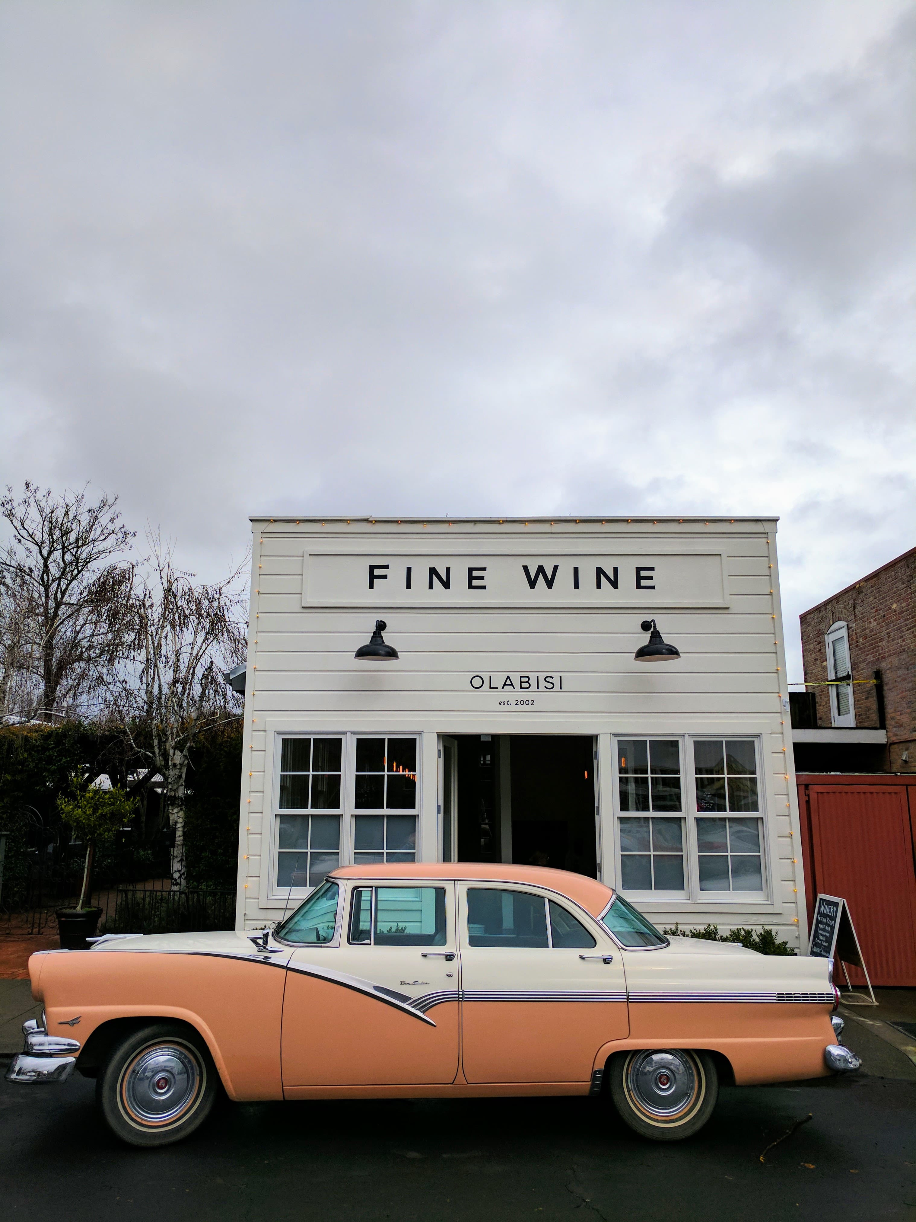 An old-fashioned orange and cream car parked in front of a white building with a sign that reads 'Fine Wine' in black lettering.