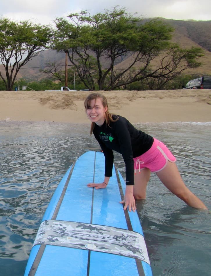 A woman posing with a blue surfboard in the shallow part of water with a beach, trees and mountains in the background.