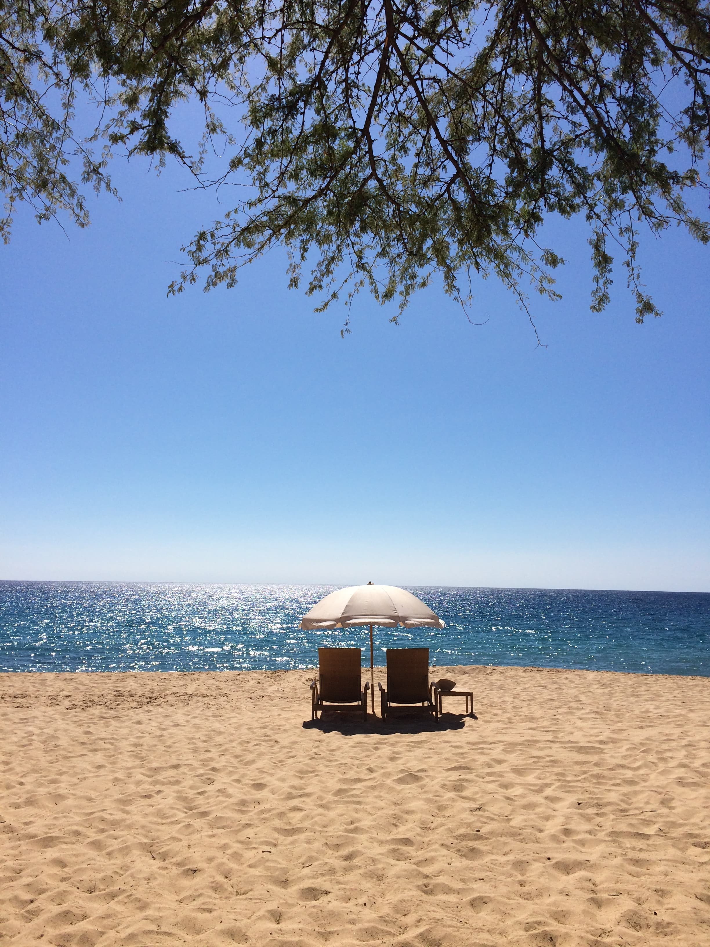 A view of a sandy beach with two lawn chairs and a white umbrella in the distance facing the sparkling blue ocean water.