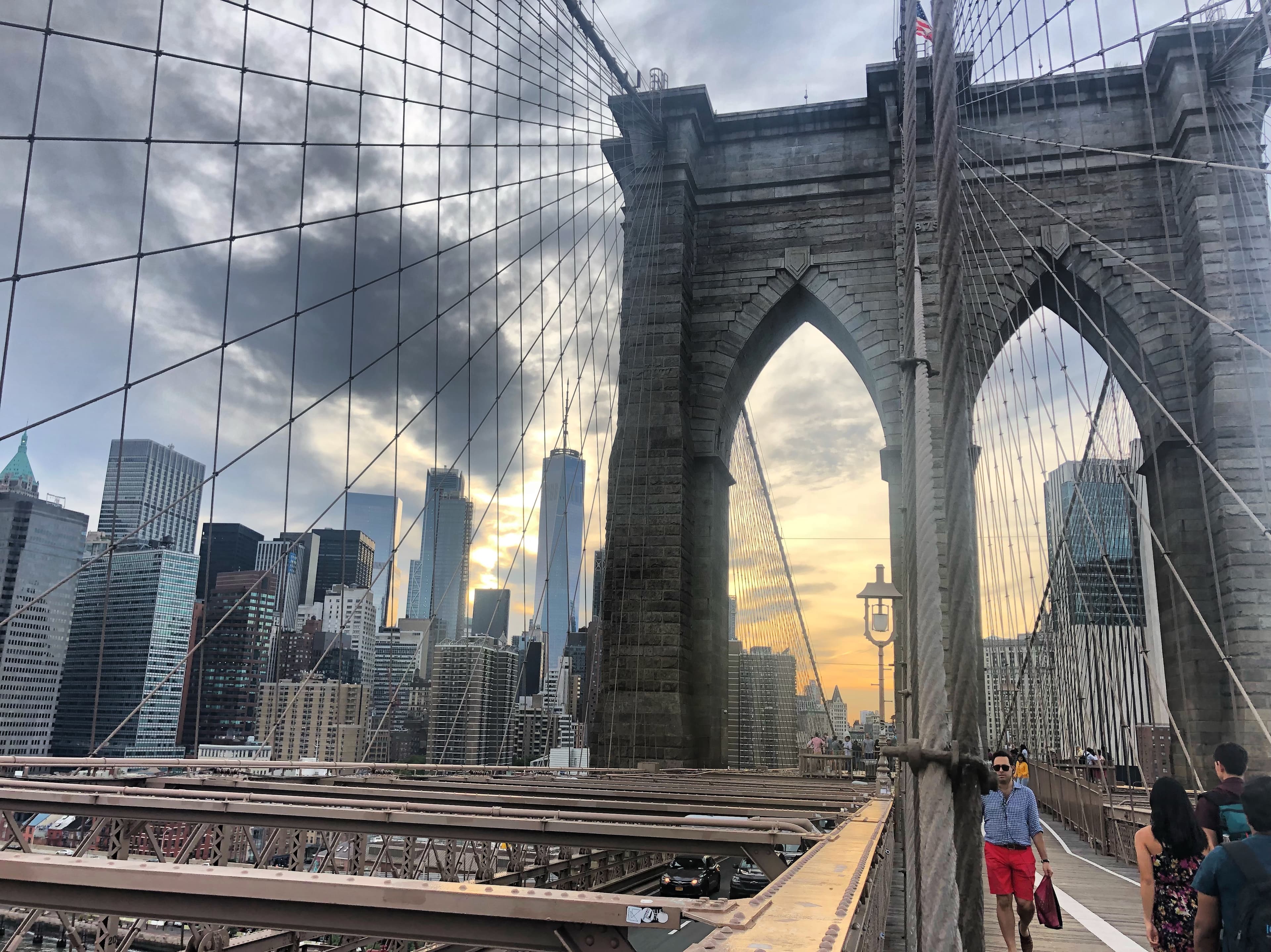 A view of the Brooklyn Bridge's beams and stone archways with the city skyline in the background and people walking in the forefront.