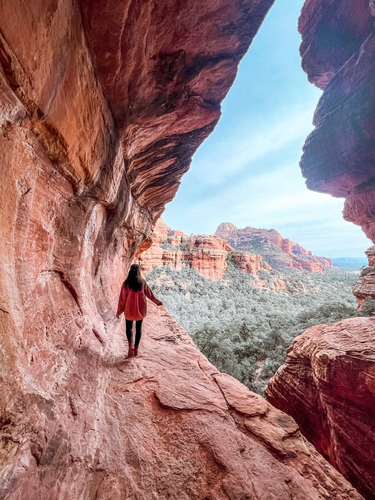 An image of Julia walking forward along canyon-like terrain with a rocky wall to her left and cliff to her right. There are trees and more canyon-like formations in the distance.