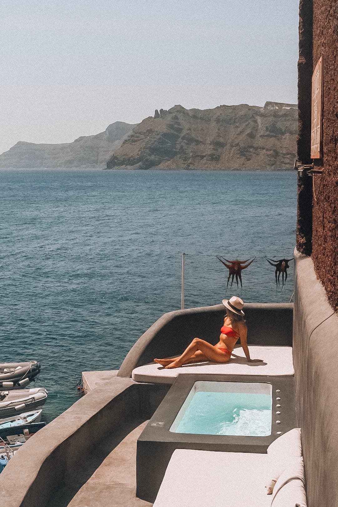 An image of Julia posing on a pool deck while wearing a straw hat and orange bikini. She is laying next to a light blue pool, stone ledge and white couch with the blue sea and mountains in the background.