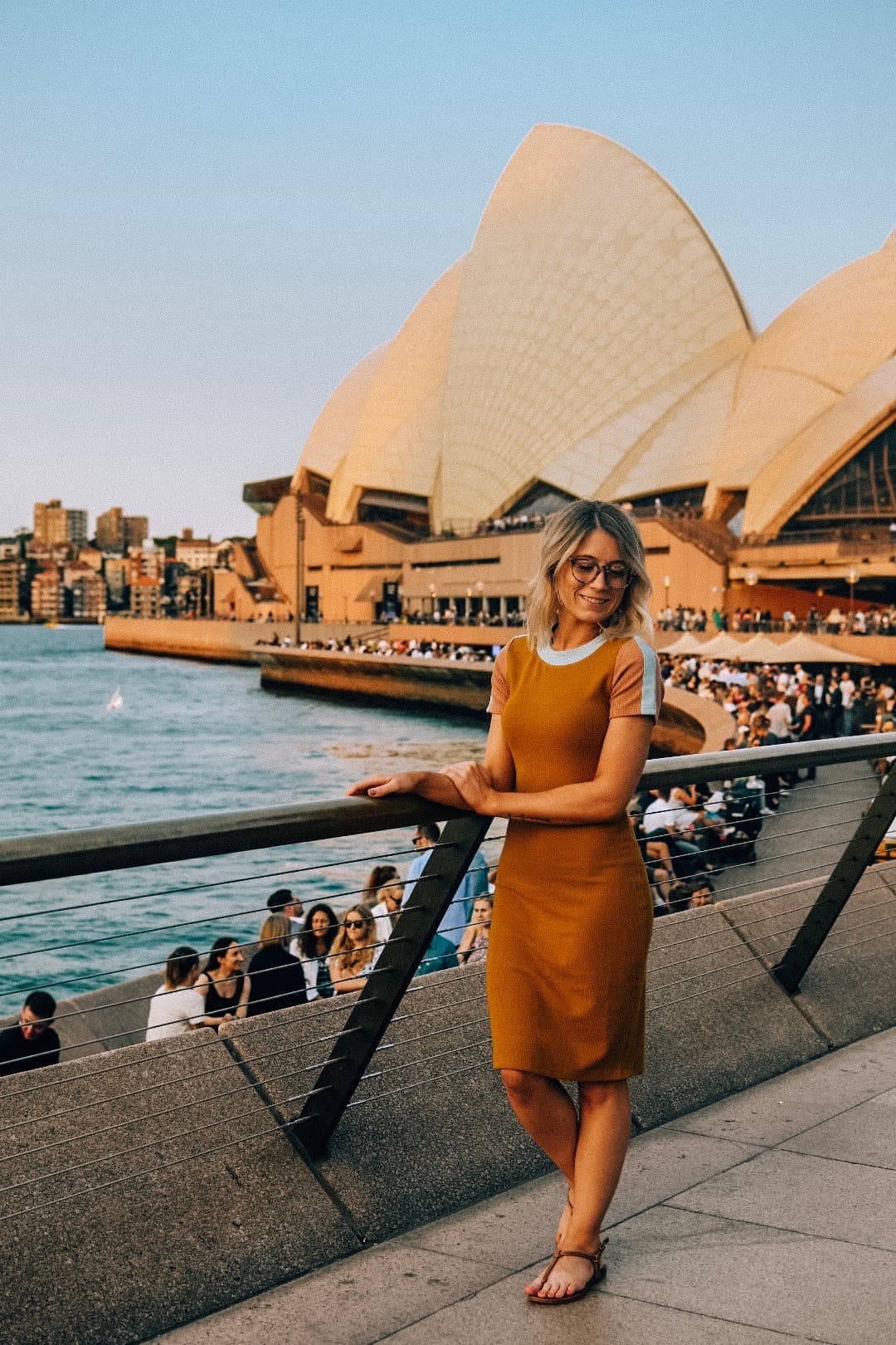 An image of Julie wearing a brown dress and posing against a metal railing in front of the Sydney Harbour and Sydney Opera house at sunset.