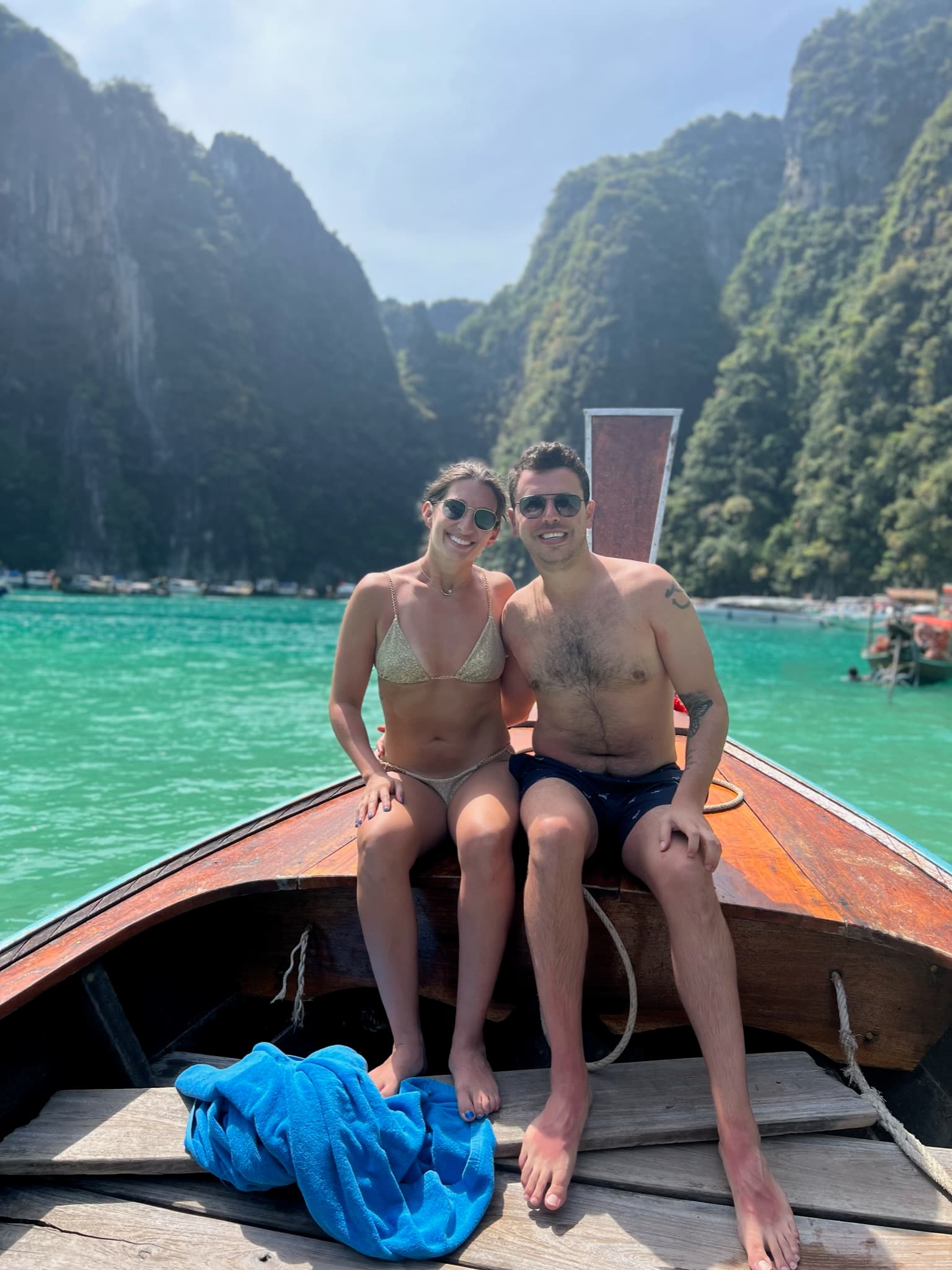Alexi and partner sitting on a wooden boat with turquoise blue water in the background and green, lush mountains.