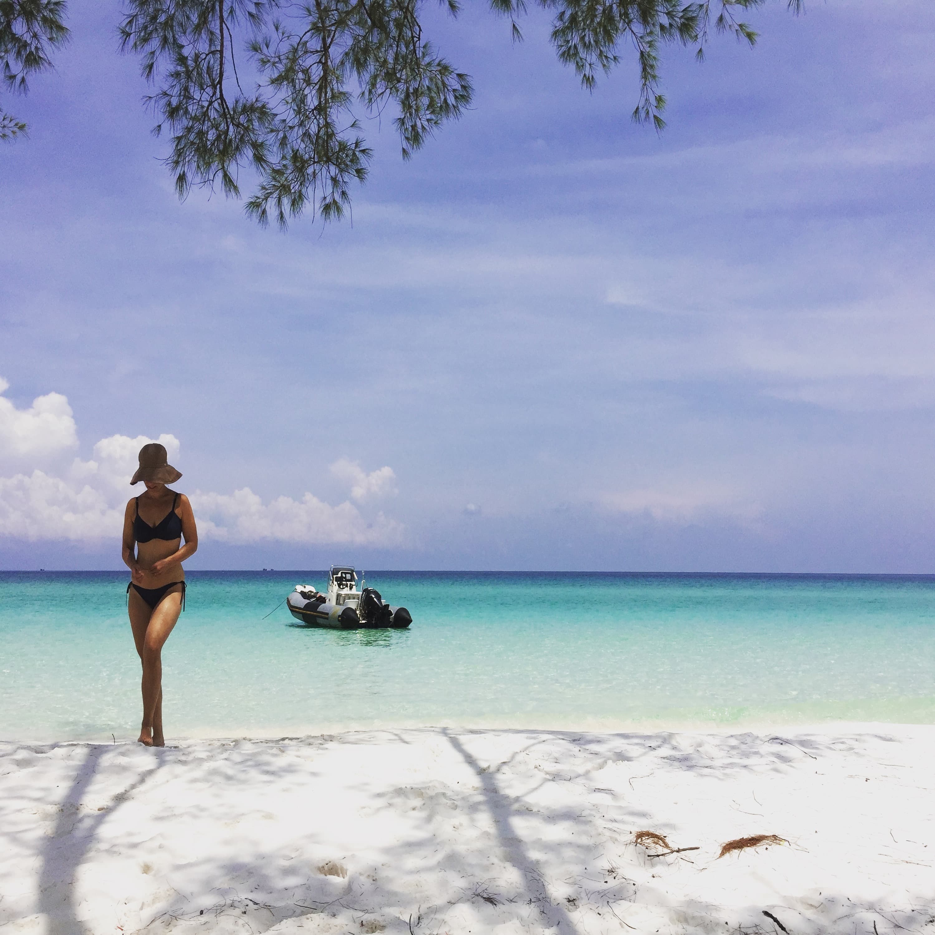 Sara standing in a swimsuit on a sandy beach with blue water and a boat in the background