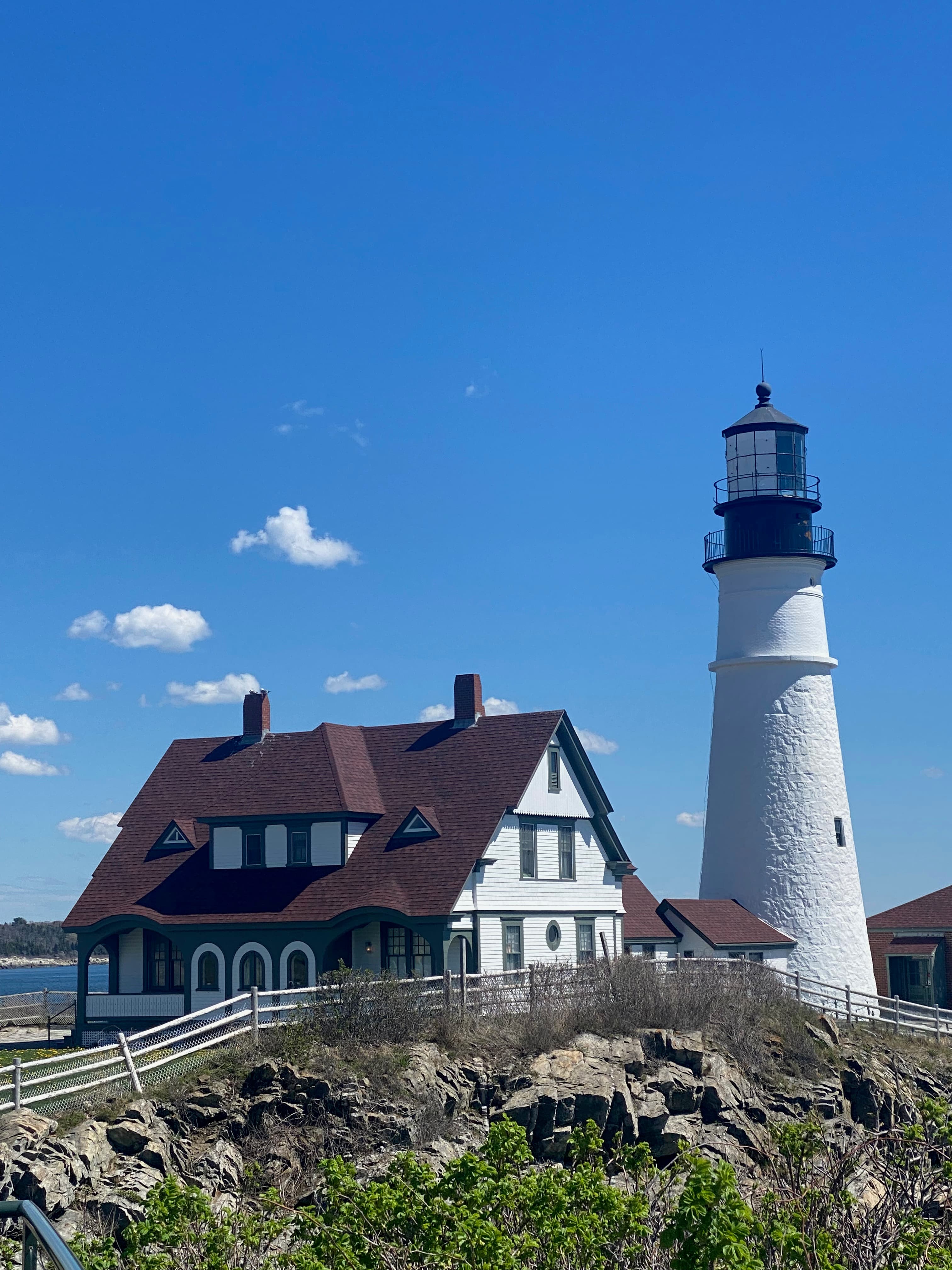 A white house and lighthouse next to a fence, rocks, green grass and the blue sea in the background under a blue sky.