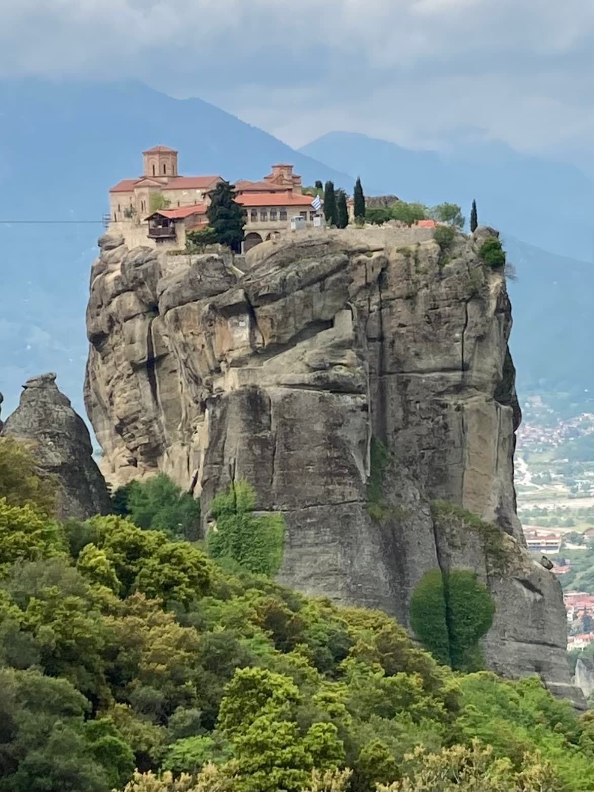 A large rock formation with a beautiful structure on top surrounded by foliage and mountains in the distance.