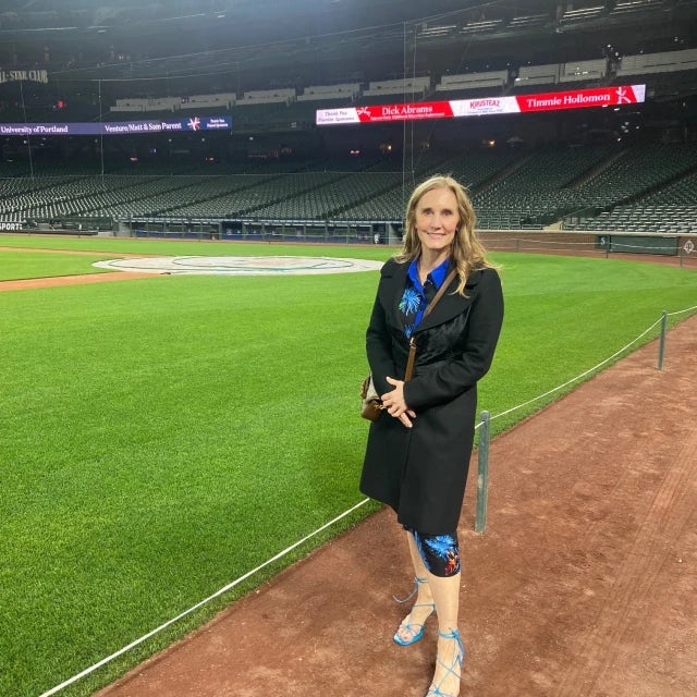 Travel Advisor Regan Hurley wears a black suit and stands on an empty baseball stadium field 