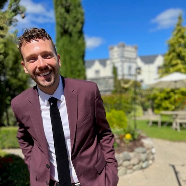 Travel advisor Cody Kaifes wears a maroon suit and black tie in a manicured garden 