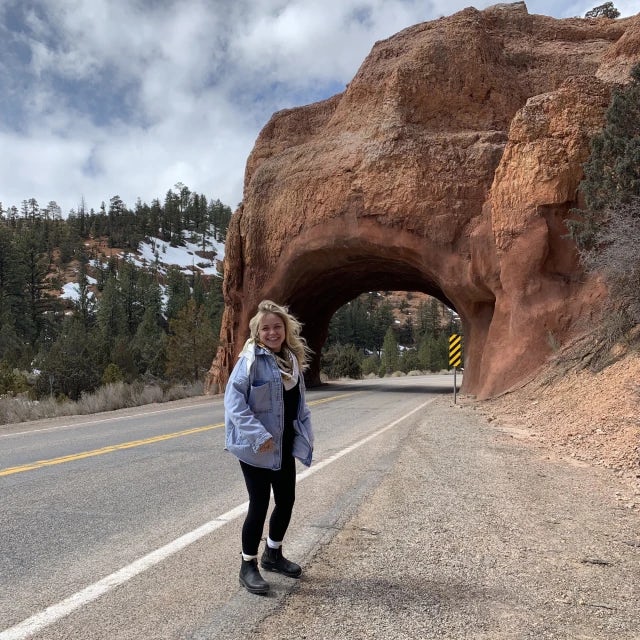 Travel Advisor Jessica Fappiano stands on a road in front of a natural stone arch wearing a jean jacket and hiking boots
