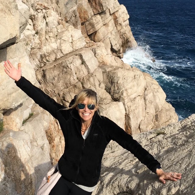 Woman wearing a black shirt and sunglasses smiles in between two craggy rocks overlooking the ocean.
