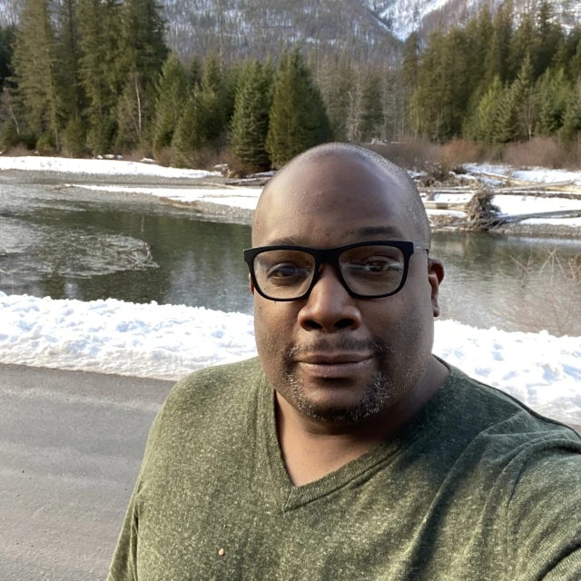 Travel Advisor Brian Hooper in a green sweater in front of green trees and snow capped mountains.