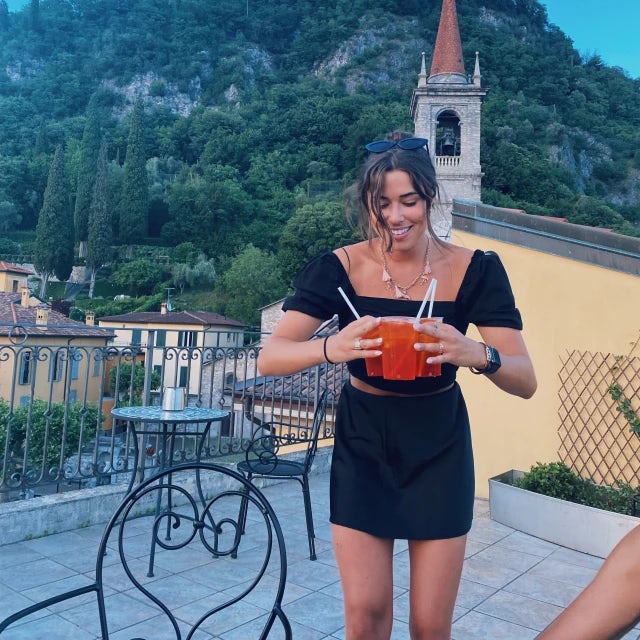 Travel Advisor Darcy Neier with drinks in her hand and a black dress in front of a yellow church and green, lush mountain.