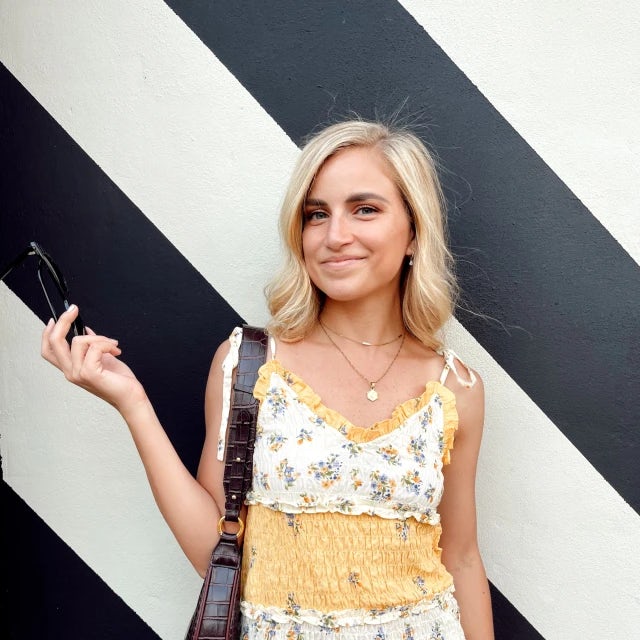 travel advisor Jasmine Masri stands in front of a black and white striped wall wearing a floral striped yellow and white sundress holding out her sunglasses