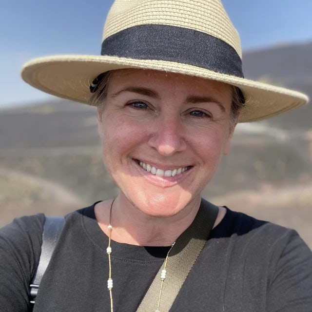 Travel Advisor Lori Loper in a black shirt and tan/black hat in front of the hills.