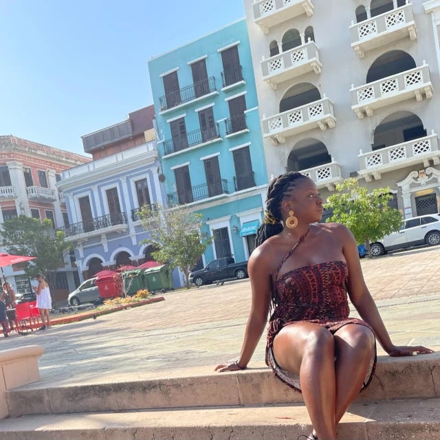 Travel Advisor Malinda Smith sitting outside in front of blue and white buildings with a red and black tube top.