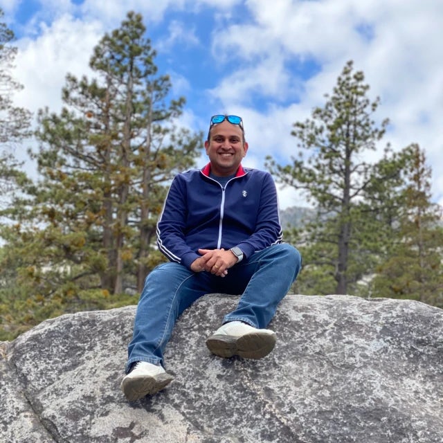 Travel Advisor Mihir Parmar with a blue jacket and jeans, sitting on a rock with trees in the distance.