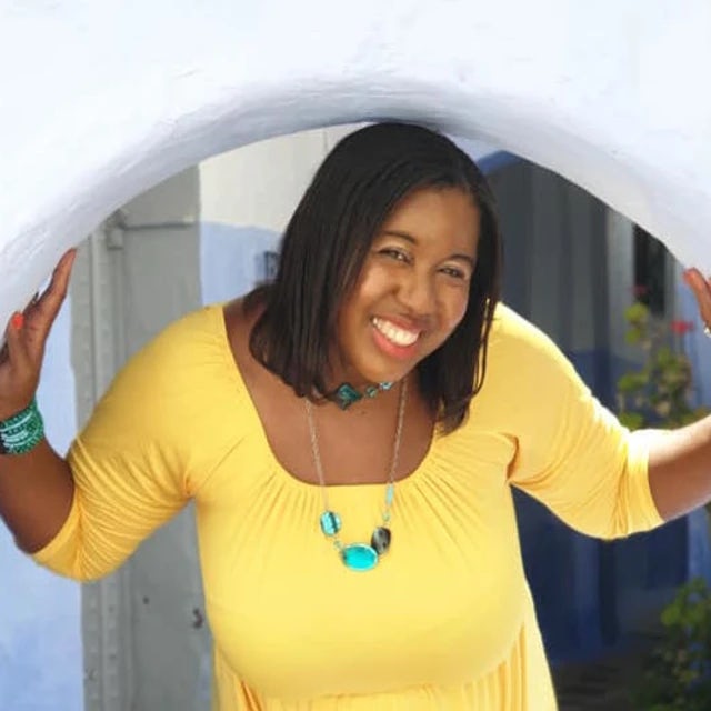 travel advisor Nicole Brewer wears a yellow top and turquoise jewelry and peaks through an arched window in a white wall