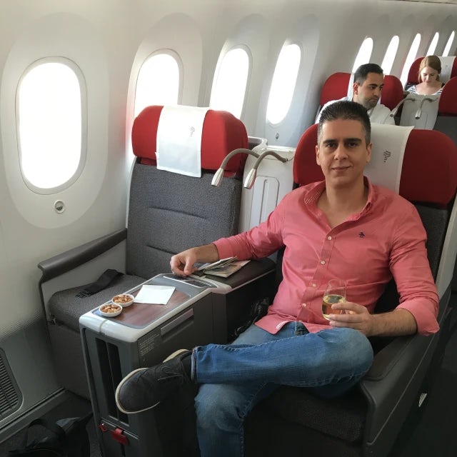 Fora travel agent Boris BK wearing red shirt and jeans sits in airplane seat