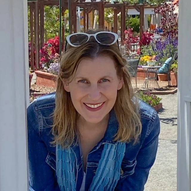 Fora travel agent Cara Todd wearing jean jacket, blue scarf and white sunglasses smiles with flowers in the background